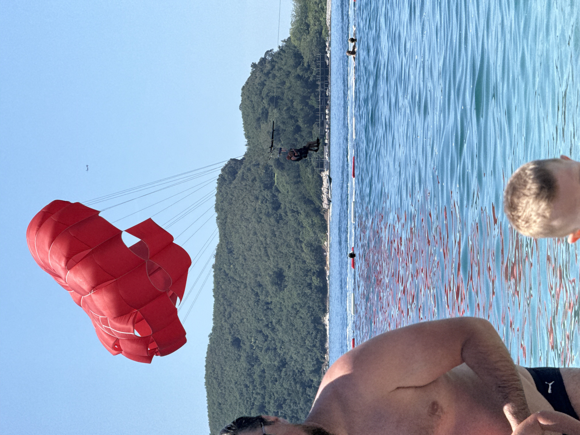 This image depicts a sunny coastal scene in Budva, Montenegro. In the upper left quadrant, a large, partially inflated red parasail dominates the sky, with numerous suspension lines extending downwards. Below the parasail, a single individual is visible, suspended by a harness system over the clear, blue water.
The background features a large, densely forested hill rising from the coastline. Along the distant shoreline at the base of the hill, several white structures, possibly buildings or docks, and small boats are visible. The water in the mid-ground is a vivid blue with surface ripples and reflections, and a line of red and white buoys marks a swimming boundary. Beyond these buoys, several people are visible in the water, swimming or wading. High in the clear blue sky, a small dark aircraft is present to the right of the parasail.
In the immediate foreground, to the left, the right shoulder, upper arm, and chest of a shirtless adult male are visible, wearing dark swim trunks with a white Puma logo. His partial face, including a beard and glasses, is visible at the top left edge. To the lower center-right, the top of a child's head with light brown hair is visible, also in the water.