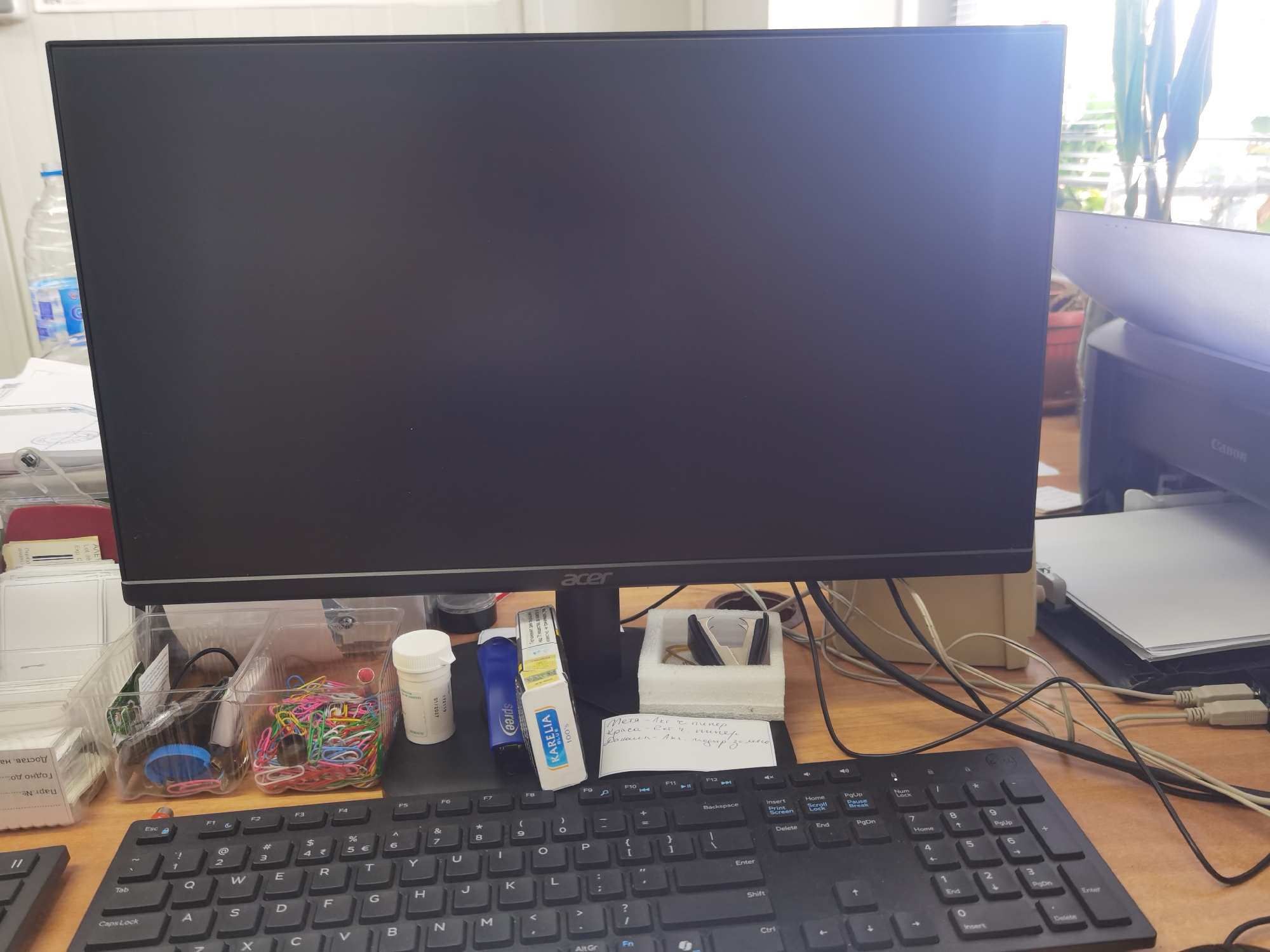 This is a forensic, matter-of-fact description of a desk scene. The main subject is a black computer monitor with the Acer logo visible on the stand. To the left of the monitor is a clear plastic container filled with colorful paper clips, and another section of the container holds a blue cap and some wires. To the right of the paper clips is a white pill bottle with a blue cap, and next to it, a blue and white package labeled "KARELIA BLUE 100%." Behind the pill bottle and Karelia package is a small white notepad with handwritten text in Cyrillic. A black keyboard with Cyrillic characters is positioned in front of the monitor. In the background, to the right of the monitor, a white printer is partially visible, with paper resting on top of it. A window with blinds and greenery is visible behind the printer and monitor, suggesting an indoor setting. The overall scene is an organized office desk. The location is not specified in the image or provided context.