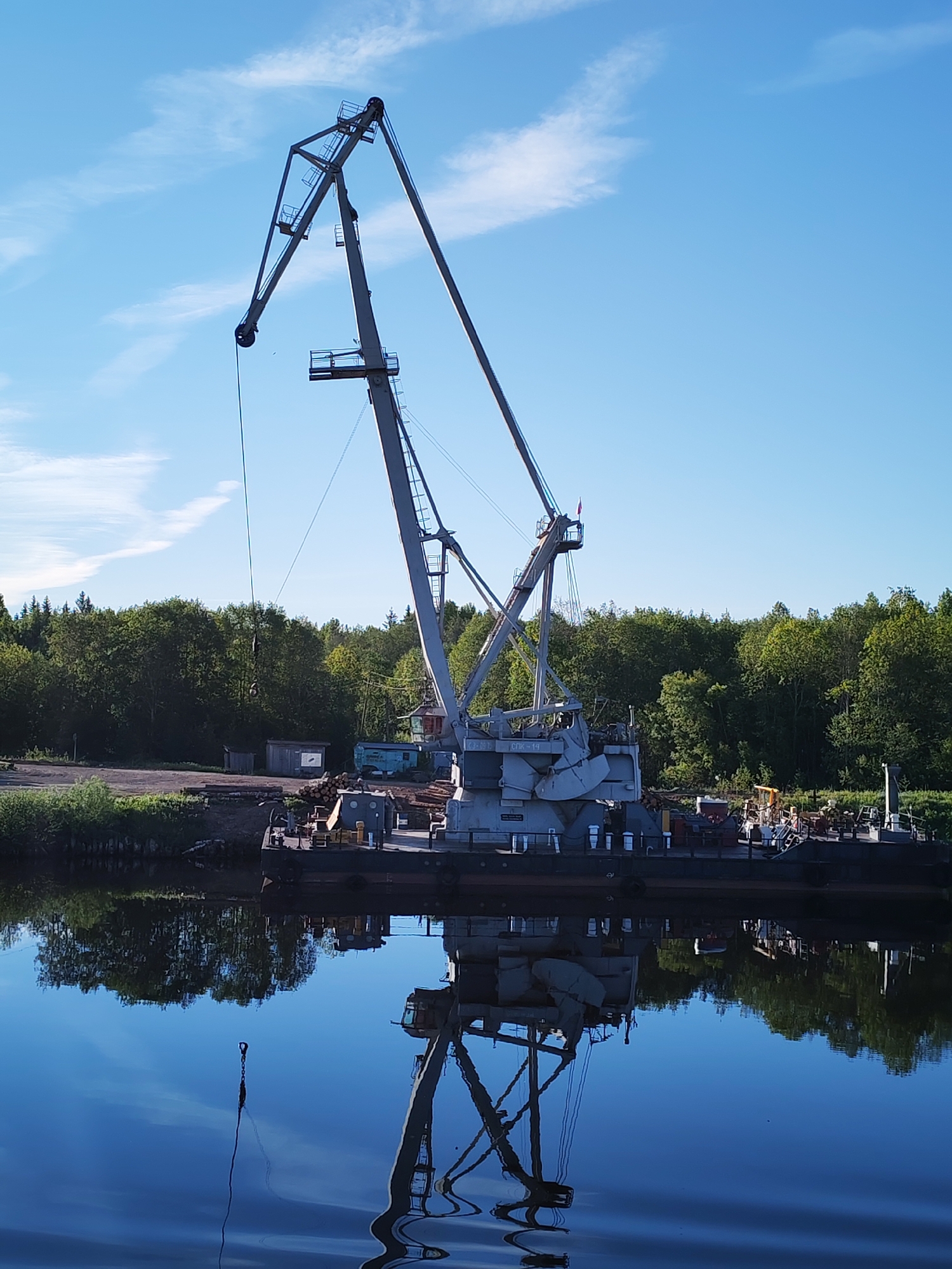 The image depicts a large industrial crane situated at the edge of a calm body of water, likely a river or canal, in Annensky Most, Russia.
The primary subject is a tall, grey industrial crane with a long, angular boom extending diagonally upwards towards the center-left. It appears to be a port or cargo crane, possibly mounted on a dark-colored barge or a fixed platform. Ladders and maintenance platforms are visible along its structure. A heavy-duty hook hangs from a cable that descends from the crane's boom, nearly reaching the water. White Cyrillic text, "СОК-18", is visible on the crane's main body near its base.
The water in the foreground provides a near-perfect, mirror-like reflection of the crane, the bright blue sky with scattered white clouds, and the lush green trees lining the banks. The stillness of the water creates a serene visual effect, with only very subtle ripples disturbing the reflection.
On the land behind the crane, there's a clearing with stacks of timber (logs), indicating a logging or lumber processing and loading operation. Several small, simple buildings are scattered amidst the trees. The background is dominated by a dense forest of various green trees.
The scene is captured in bright daylight, suggesting late morning or early afternoon on a clear, sunny day. The weather appears fair. No human figures are visible, and the crane appears to be idle, suggesting a break in activity at this industrial waterside location.