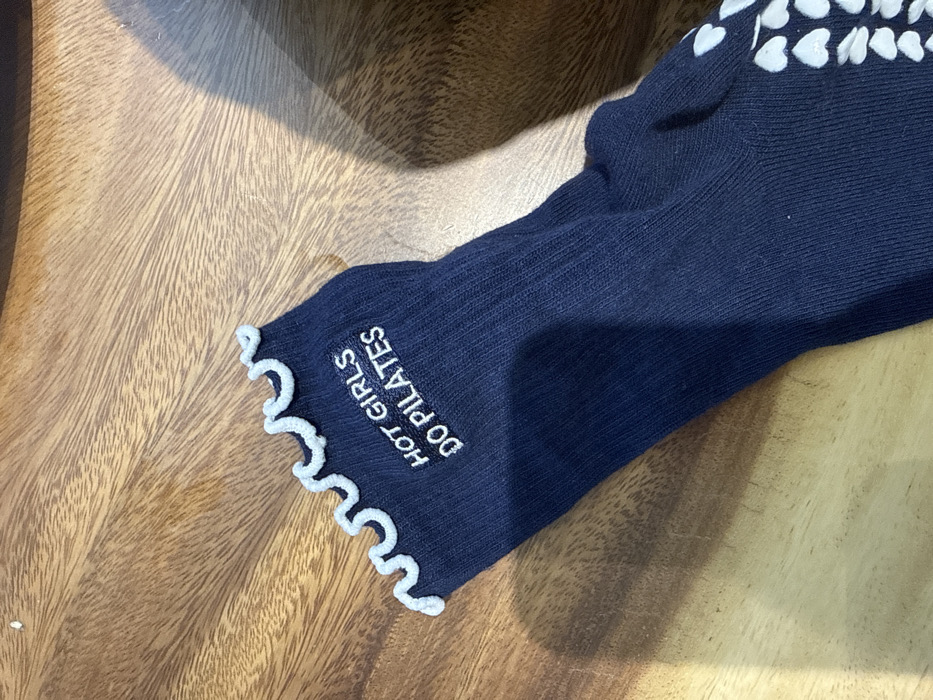 A single navy blue, ribbed sock is depicted resting on a wooden surface in Dubai, United Arab Emirates. The sock features a light blue ruffled cuff at its opening. White embroidered text on the upper portion of the sock reads "HOT GIRLS DO PILATES". The lower section of the sock, partially visible, displays multiple light blue heart-shaped grip dots. The background consists of a medium-brown wooden surface with visible grain patterns, partially shadowed on the right side of the frame. No individuals are present in the image.