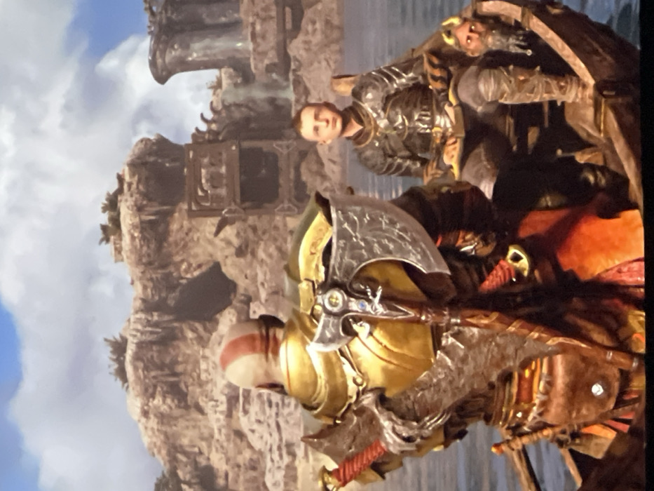 The image shows Kratos and Atreus in a boat, likely from the video game "God of War." Kratos is in the foreground, facing left, with his Leviathan Axe prominently displayed. He wears golden armor. Atreus stands behind him, gazing directly at the viewer, wearing similar armor. The background reveals a rocky shoreline and a partially visible building or structure. The sky is cloudy.