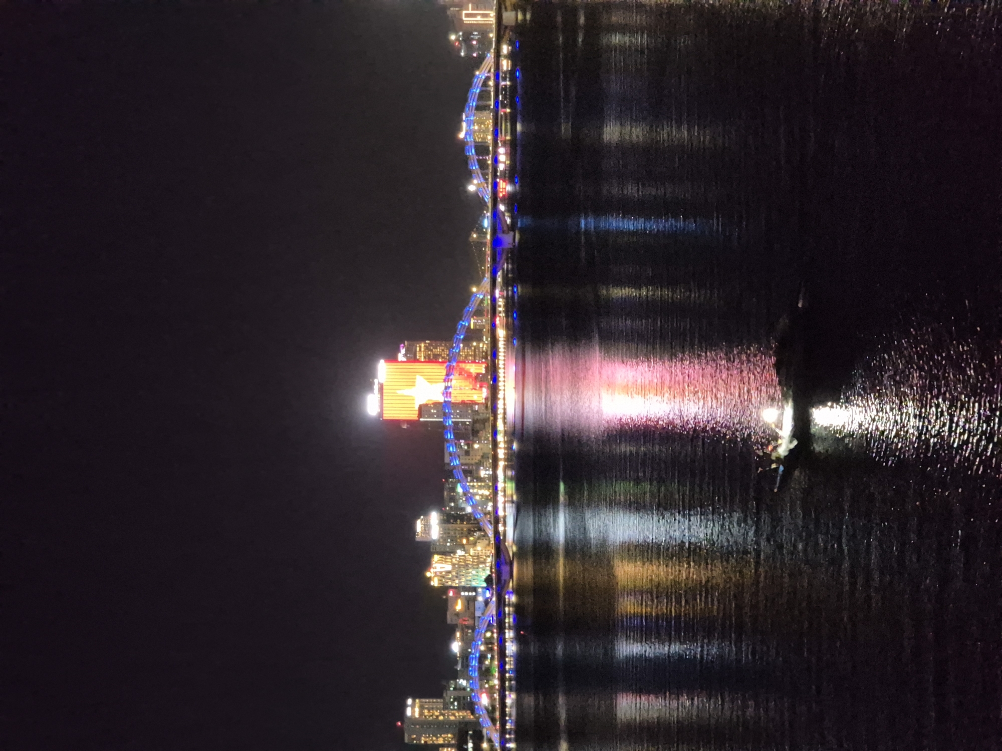 This image captures a nighttime view of the Dragon Bridge in Da Nang, Vietnam, spanning the Han River. The bridge is illuminated with blue lights, creating a striking visual against the dark sky. To the right of the bridge, a tall building is prominently lit with a large, yellow star emblem against a red background. The cityscape along the riverbank features several high-rise buildings, all aglow with various colored lights. The river itself reflects the vibrant city lights, creating a shimmering effect on the water's surface. In the foreground, a small boat with a light on its stern is navigating the water. The overall scene depicts a lively urban environment at night, with the illuminated bridge and buildings being the focal points.