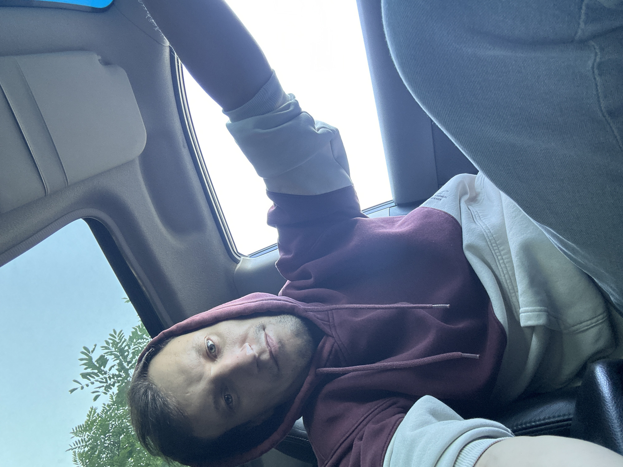 A medium shot captures a man inside a vehicle, looking towards the camera with a neutral expression. He is wearing a maroon hoodie with cream-colored sleeves and light blue jeans. The man's right arm is resting on what appears to be the car's interior door panel, with his elbow bent and hand visible. The vehicle's sunroof is open, allowing daylight and a glimpse of green foliage and the sky overhead. The interior of the car, including a sun visor and part of the dashboard, is also visible. The lighting suggests it is daytime. The image is a selfie, taken from a slightly low angle.