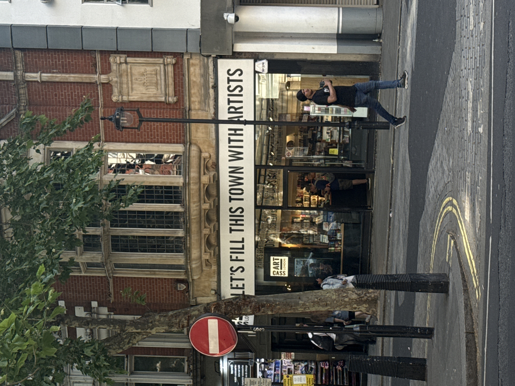 Here is a detailed description of the image:
The image depicts a street scene. The building on the left is of a red-brick architecture style with multiple windows and decorative architectural elements. Next to it, is a shop front, decorated with a white banner reading "LET'S FILL THIS TOWN WITH ARTISTS". The shop's window displays various art supplies and posters.
On the sidewalk next to the shop, there's a man taking pictures with a camera. A "No Entry" sign is visible, attached to a pole beside a tree. Another pedestrian is visible closer to the camera.
The road surface is asphalt, marked with yellow lines near the curb. The composition suggests an urban environment.