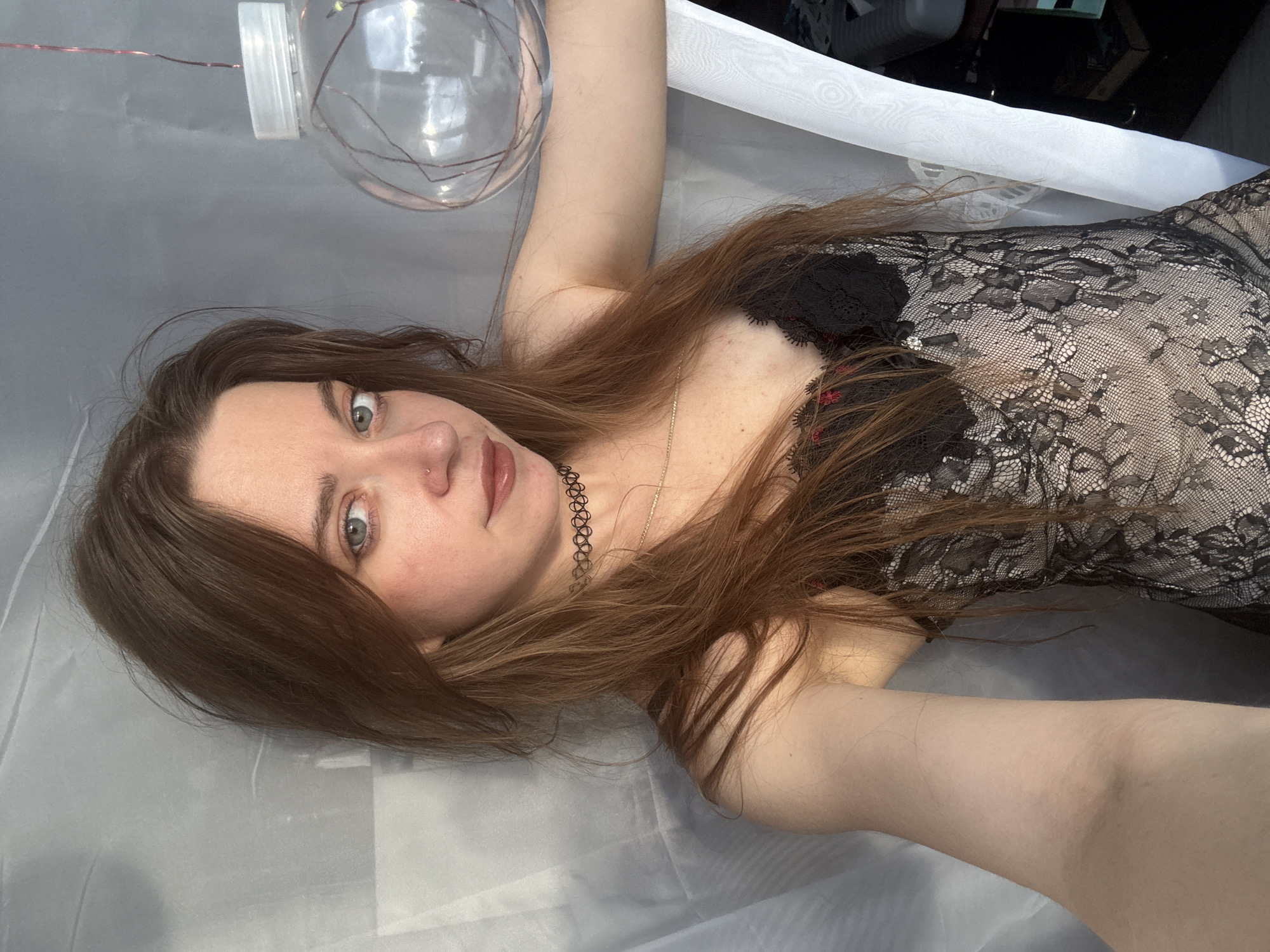 A young woman with long, light brown hair and blue eyes is pictured from the chest up, looking directly at the camera. She is wearing a black lace top with a delicate choker necklace. Her head is tilted slightly to her right, and she is positioned in front of a sheer white curtain. To her right, a clear glass orb filled with string lights is visible. The lighting suggests it is daytime, with natural light filtering in. The background is a soft grey.