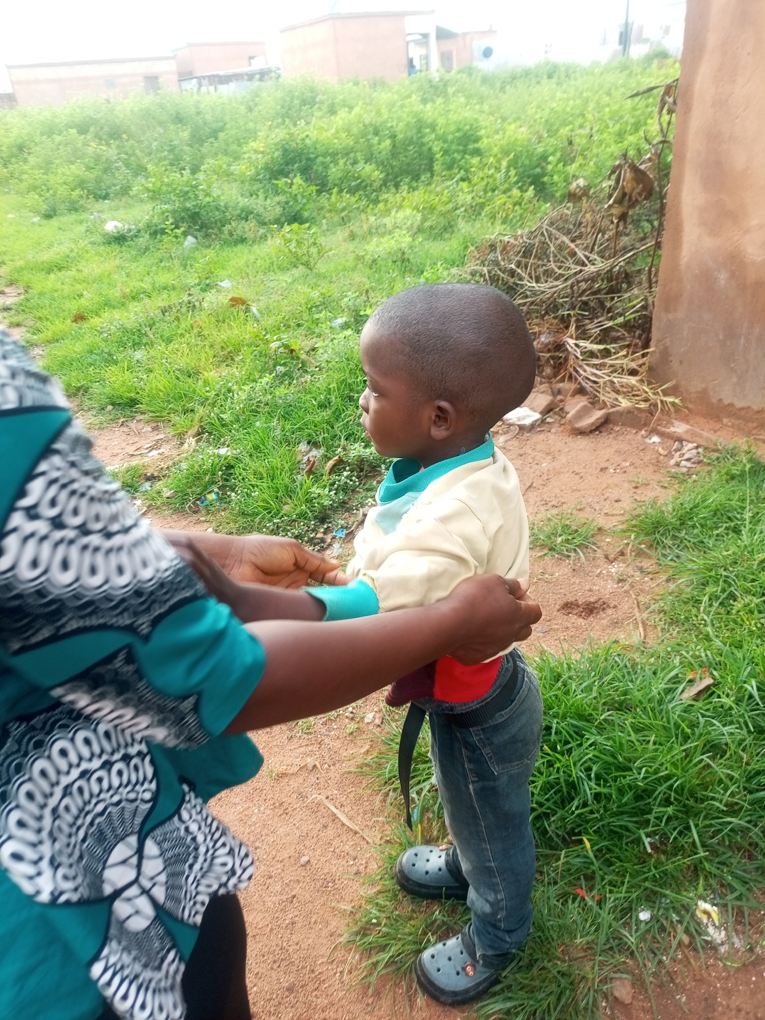 The image shows a young child standing outdoors, facing to the right. The child has short, dark hair and is wearing a light-colored long-sleeved shirt with a turquoise collar and cuffs, and blue jeans. The child's feet are in grey clogs. A person, whose body is partially visible on the left of the frame, is holding the child by the waist. This person is wearing a blue and white patterned garment.
The setting appears to be a rural or semi-rural area. In the background, there is green vegetation, possibly grass and bushes, and some buildings. The ground is a mix of dirt and grass. The lighting suggests it is daytime, and the overall atmosphere seems casual. There are no visible signs of text in the image. The location context provided is Tempure, Nigeria.