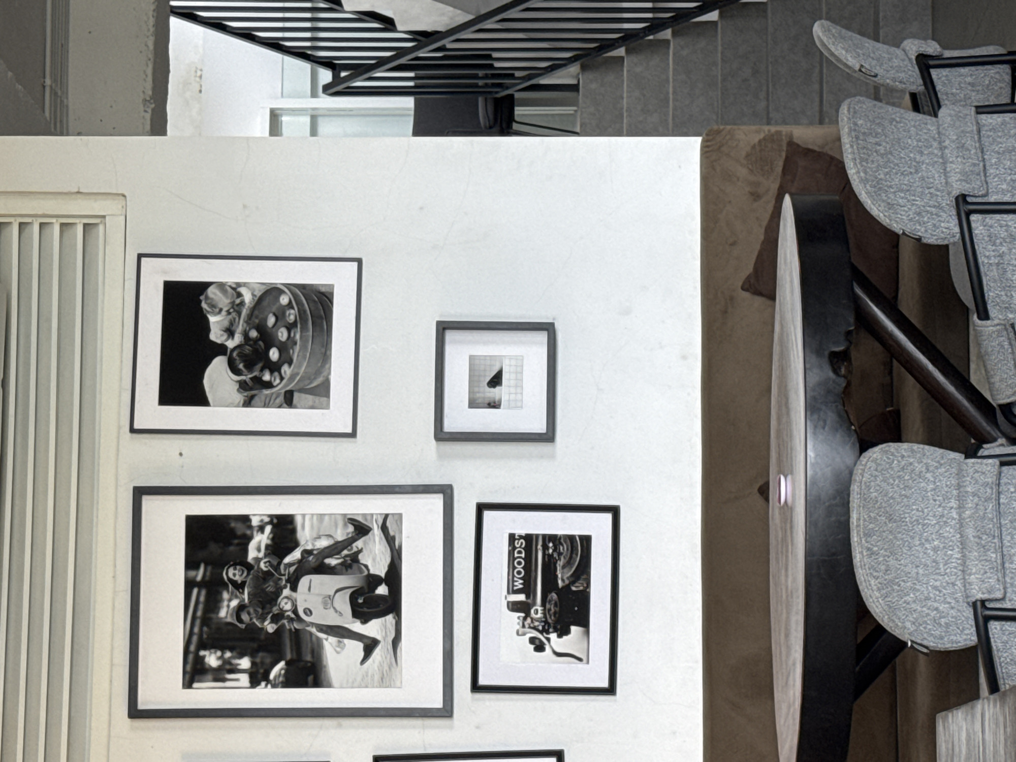 This is an interior scene, featuring a wall with several framed black and white photographs hanging on it. To the left of the wall is a section of vertical blinds or louvers. Above the wall, there is a glimpse of a staircase with a metal railing. To the right of the wall, there is a piece of furniture with a rounded edge and several high chairs with gray textured seats. The environment suggests a modern or urban setting. The exact location and specific activities are unknown.