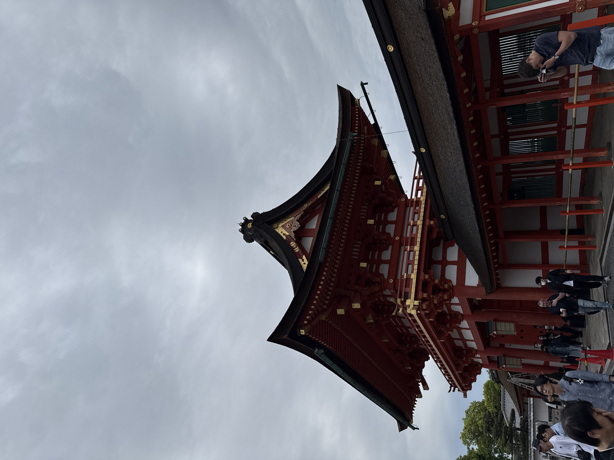 The image captures a low-angle view of the Fushimi Inari Shrine in Kyoto, Japan. The distinctive vermillion red architecture is prominently featured, with intricate details of the multi-tiered roof and support structures visible. A portion of a dark-colored thatched roof is also visible. A group of people are seen near the shrine, some wearing face masks. One person is actively taking a photograph. The sky is overcast with cloudy conditions.