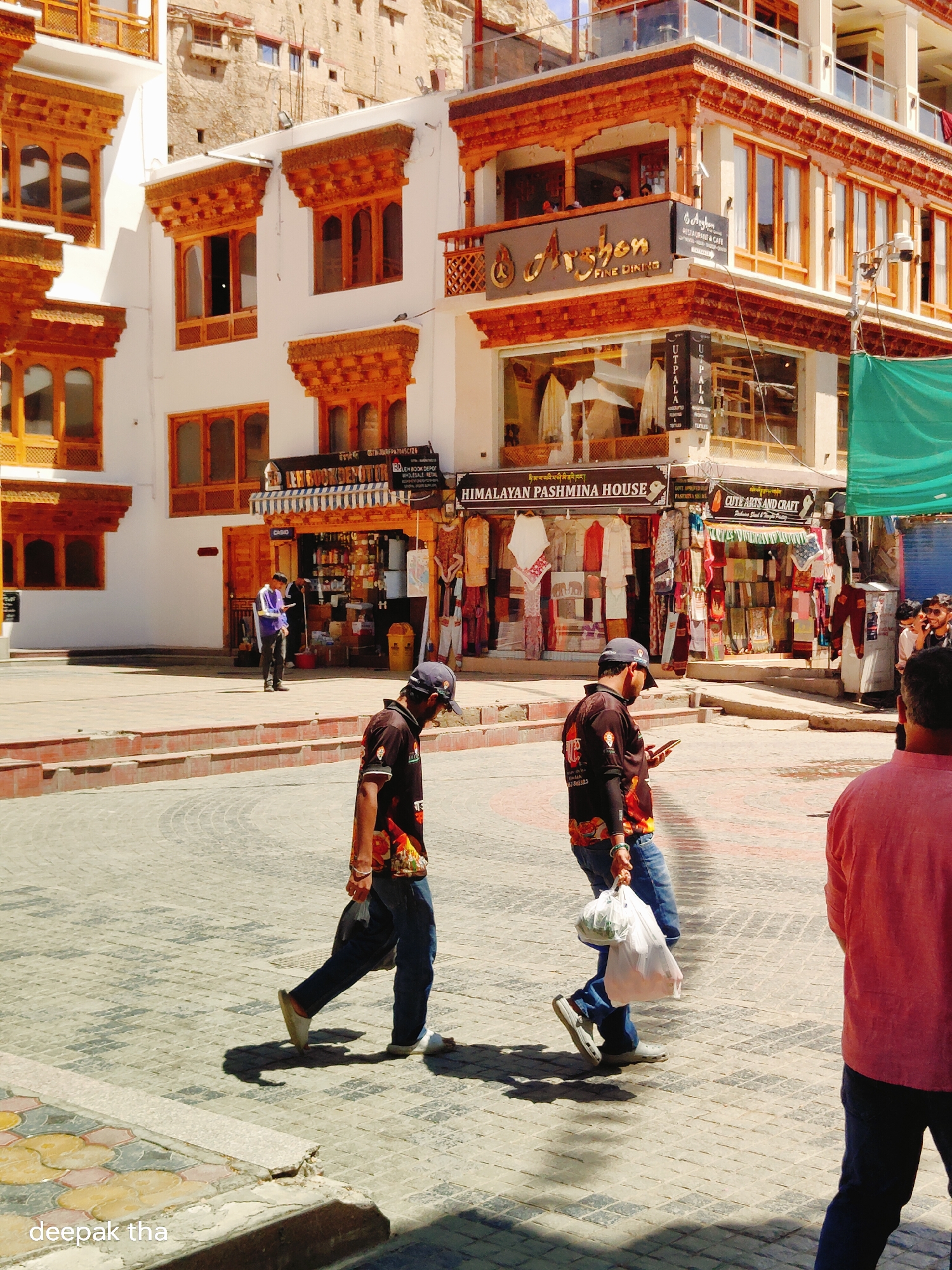 The image captures a street scene in Leh, India, with distinctive Ladakhi architecture. The buildings feature white facades adorned with intricate wooden window frames and eaves painted in warm orange hues. In the foreground, two men are walking across a paved courtyard. One man, wearing a black t-shirt with colorful graphics and blue jeans, carries a white plastic bag filled with groceries in his right hand and looks down at his phone. He is wearing a dark baseball cap. The second man, also in a t-shirt and jeans, is walking to his left, carrying a similar white plastic bag. Further back, another man in a blue jacket stands near a storefront, looking at his phone. The storefronts display various goods, including textiles and handicrafts. A green awning is visible on the right side of the image, and a security camera is mounted on a pole above one of the shops. The ground is paved with interlocking bricks. The overall atmosphere suggests a bright, sunny day in a busy marketplace.