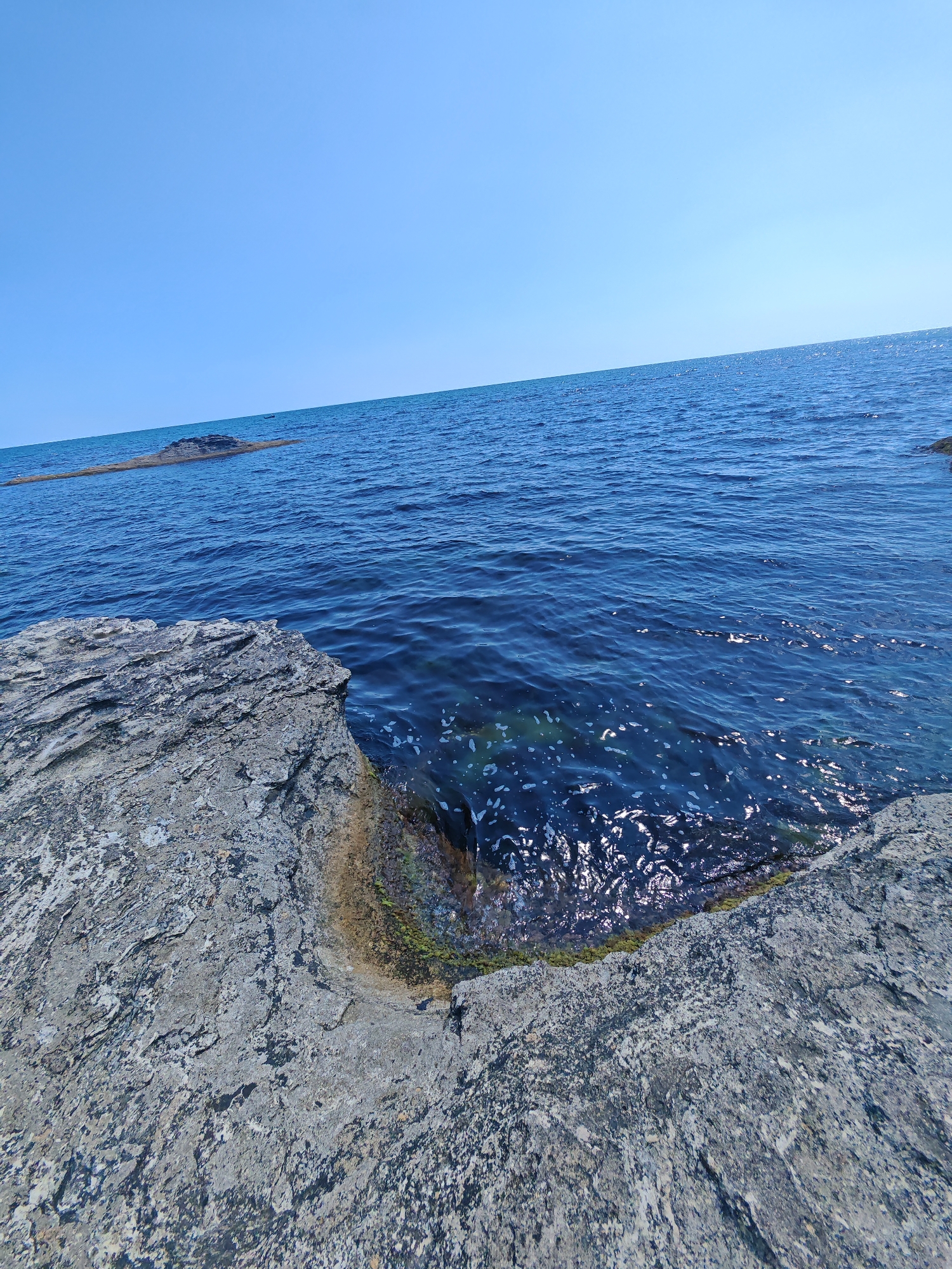 The image shows a rocky coastline on a clear day. The foreground is dominated by large, textured gray rocks that extend into the frame from the left and bottom. The sea is a deep blue, with small waves and sunlight glinting off the surface. A small, dark rock formation protrudes from the water in the distance, and the horizon line is sharp against a clear, pale blue sky. The location is the coast of Tsarevo, Bulgaria. There are no people or animals visible in the image.