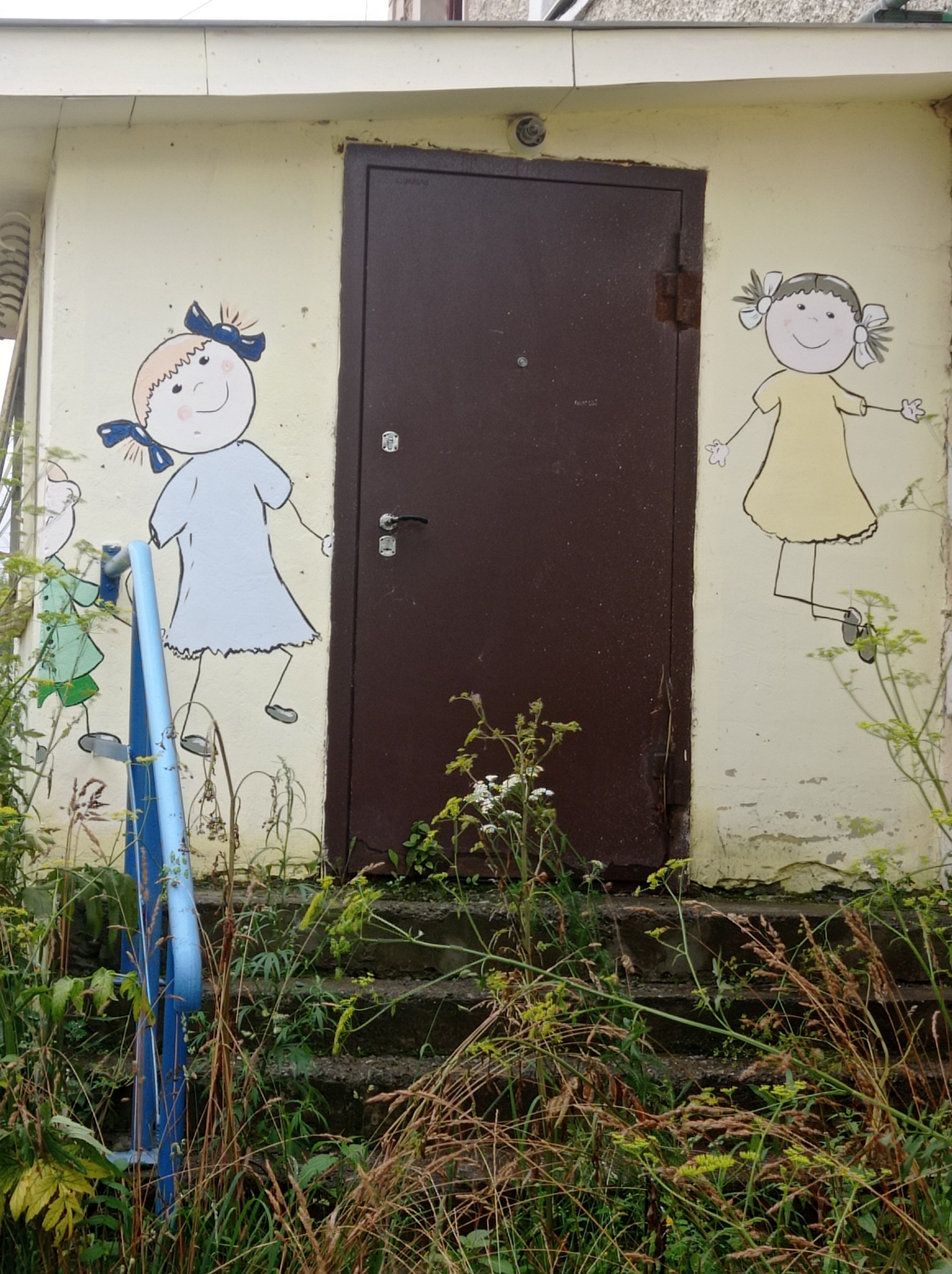 An exterior shot captures a weathered, pale yellow building with a dark brown door. On the left side of the door, a cartoon drawing of a young girl with blue bows in her hair wears a light blue dress. To the right of the door, another cartoon drawing depicts a young girl with pigtails and yellow bows in her hair, wearing a yellow dress. The building's lower section is surrounded by overgrown weeds and grass, with concrete steps leading up to the door. A blue metal railing is visible on the left side, partially obscured by the vegetation. The overall scene suggests an outdoor area in an urban or semi-urban environment, possibly a residential building or a public facility in Degtyarsk, Russia.