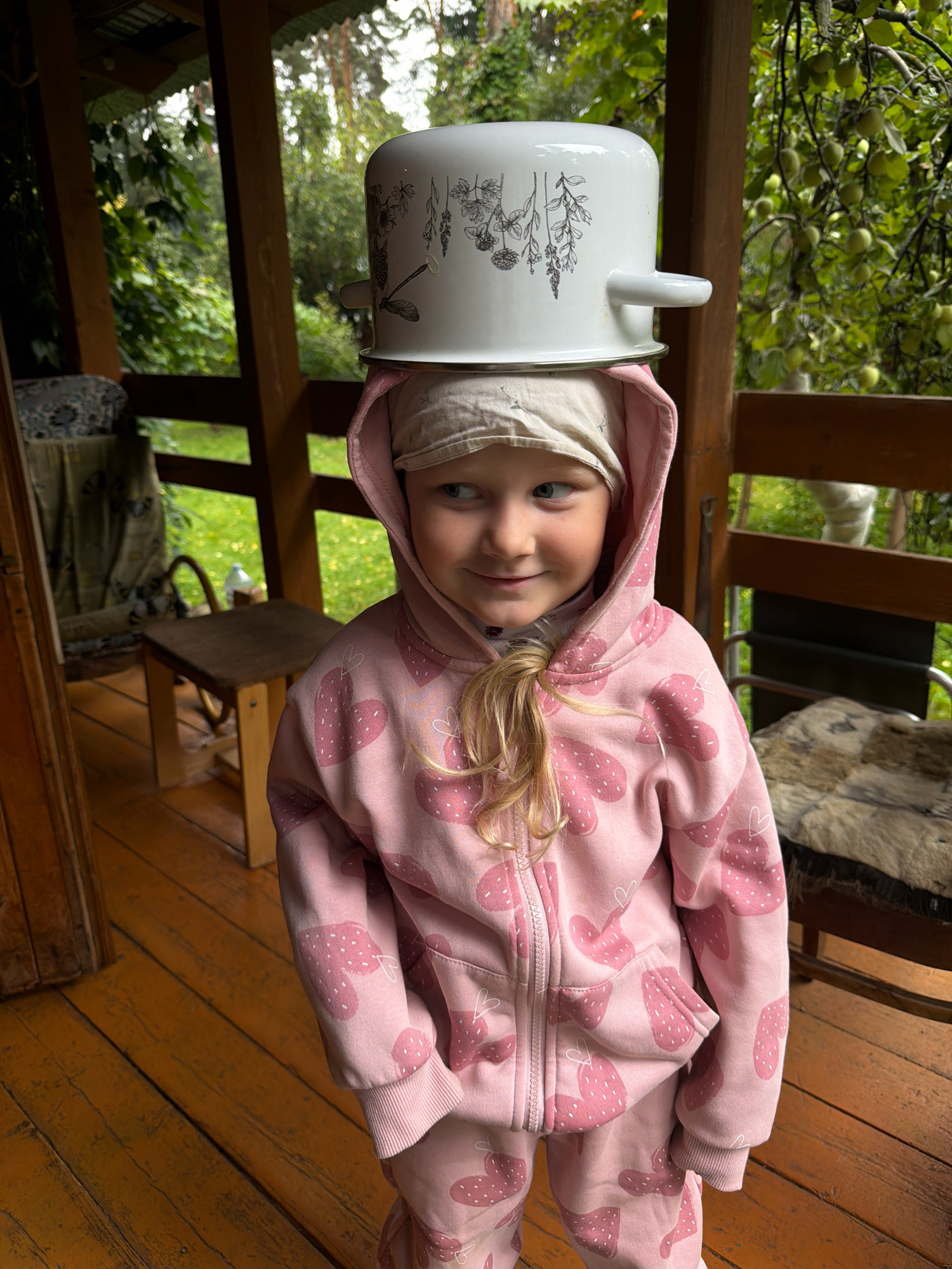 In the city of Udelnaya, Russia, a young girl is pictured standing on a wooden porch. She is wearing a pink hooded tracksuit adorned with a strawberry pattern and a beige headscarband. A white pot with botanical drawings is balanced on her head. The porch has wooden floorboards and railings, with glimpses of green trees and foliage in the background. A wooden chair is visible on the left side of the porch, and another chair with a fur covering is on the right. The lighting suggests an overcast day.
