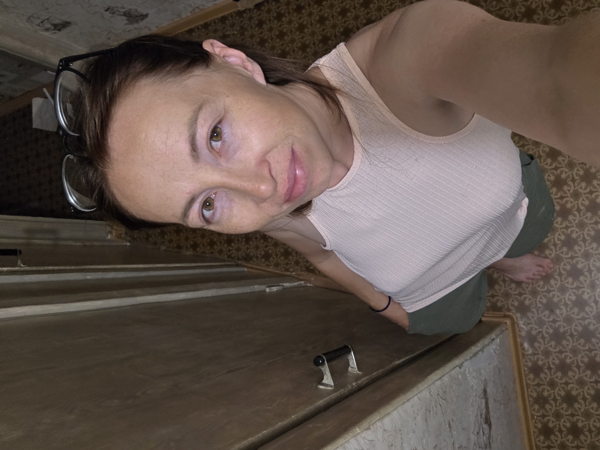 A woman with brown hair and glasses pushed up on her head is standing in a narrow indoor hallway. She is wearing a light pink ribbed tank top and green pants. She is barefoot and her left hand is in her pocket. The hallway has a dark wooden door on the left and a patterned carpet on the floor. The walls are wallpapered and appear somewhat worn. The lighting is dim.