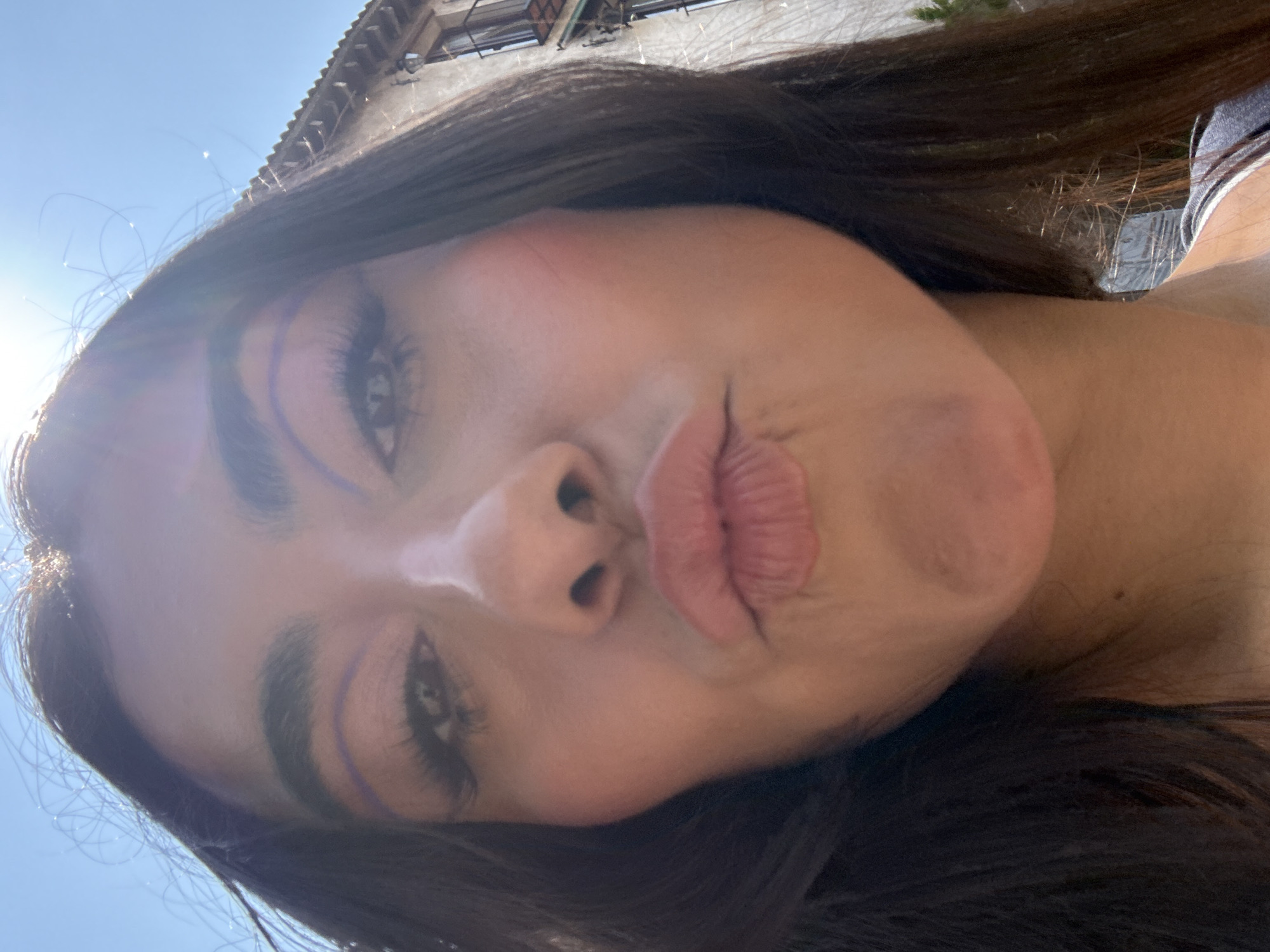 A selfie of a young woman with long dark hair, light skin, and accentuated eyes with blue eyeliner, pursing her lips. The background shows a clear blue sky with a hint of the sun's glare and part of a building with a textured roof, likely in an urban setting in Domingo Arenas, Mexico.