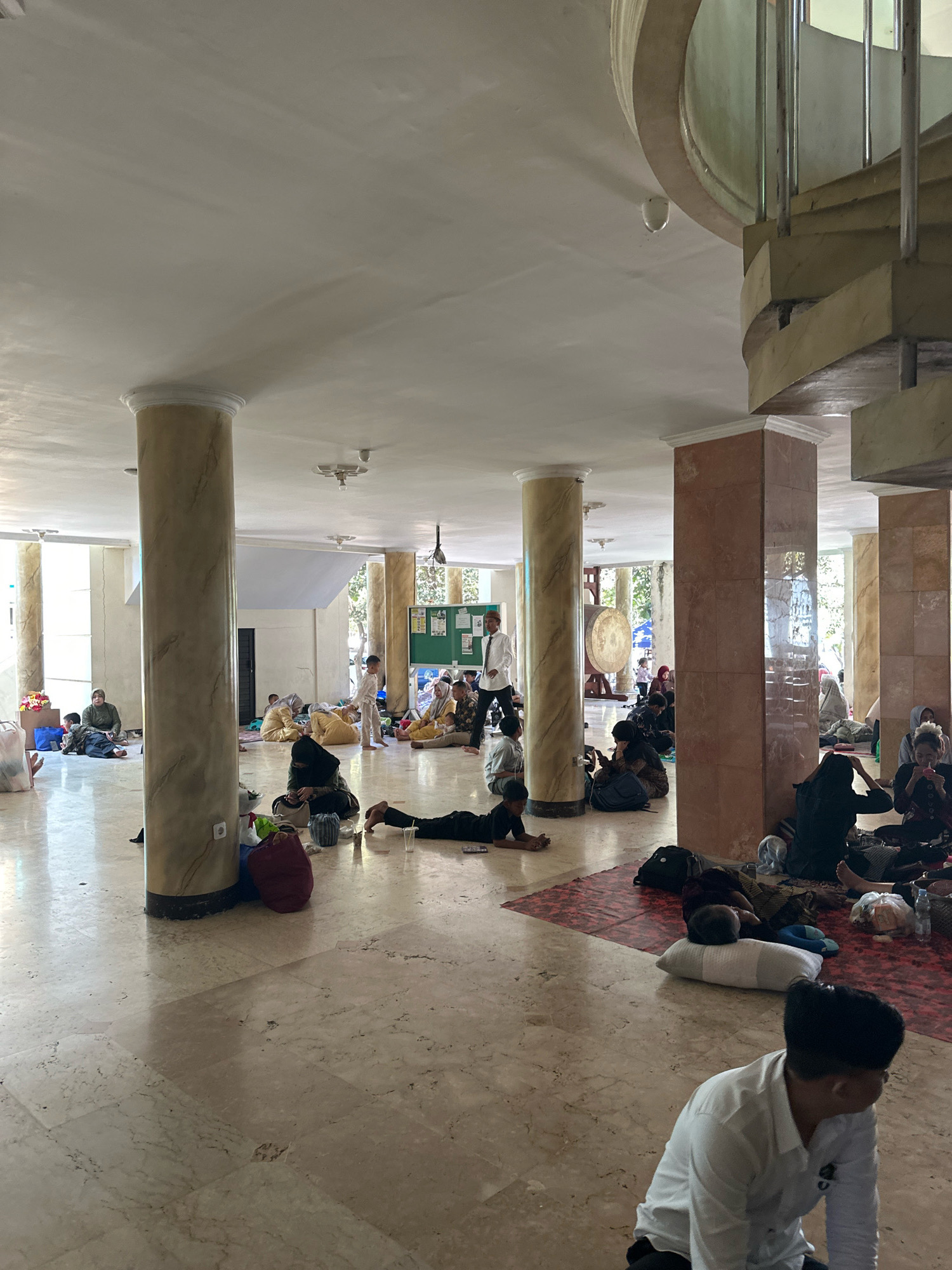 The image depicts the interior of a large, open hall, likely a community center or a mosque, filled with many people resting on the floor. The architecture features large, marble-clad pillars, a marble floor, and a high, white ceiling with ornate trim. A spiral staircase with metal railings and marble steps is visible in the background.
Dozens of individuals, appearing to be mostly women and children, are spread across the floor, many lying down or sitting with their belongings. They are dressed in casual clothing, with some wearing headscarves. Some individuals are seen with pillows and blankets, suggesting they are resting or sleeping. A large drum is positioned behind one of the pillars. The overall atmosphere appears to be one of temporary accommodation or a gathering for an event. The context provided indicates this scene is in the city of Malang, Indonesia.