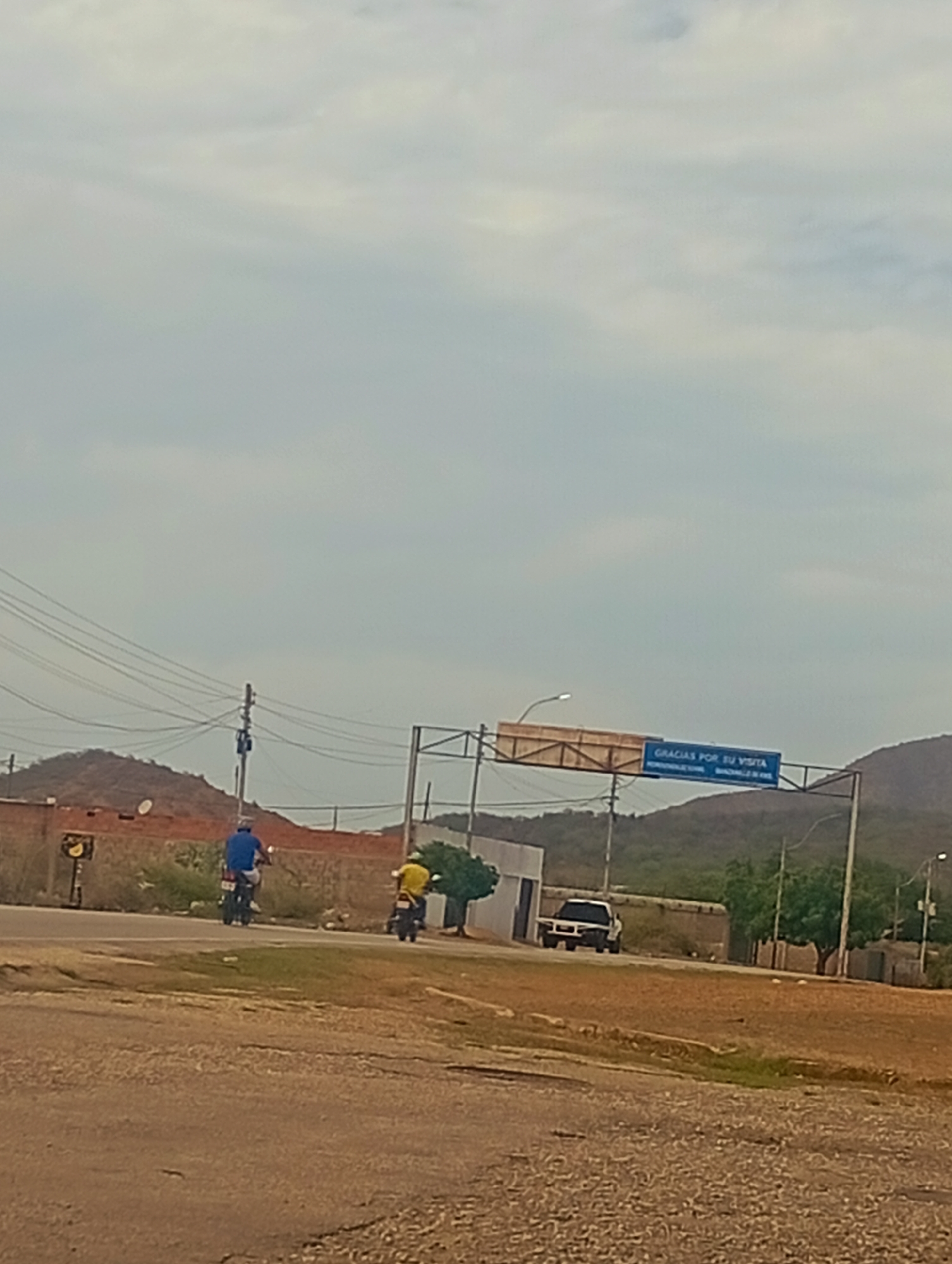 In Altagracia, Venezuela, on a cloudy day, a rural road scene unfolds. Two individuals on motorcycles are moving away from the camera on a paved road that curves to the left. The rider in the foreground wears a blue shirt and is on a dark motorcycle. Behind him, a second individual in a yellow shirt rides a dark motorcycle. To the left of the road, a brick building stands with a traffic signal pole adjacent to it. Further in the background, hills covered in vegetation are visible under a hazy sky. An overhead sign with blue lettering stating "GRACIAS POR SU VISITA" (Thank you for your visit) is suspended over the road. A white pickup truck is parked on the right side of the road near a cluster of trees. The ground in the foreground is rough and unpaved, transitioning to dry grass and dirt on the sides of the road.