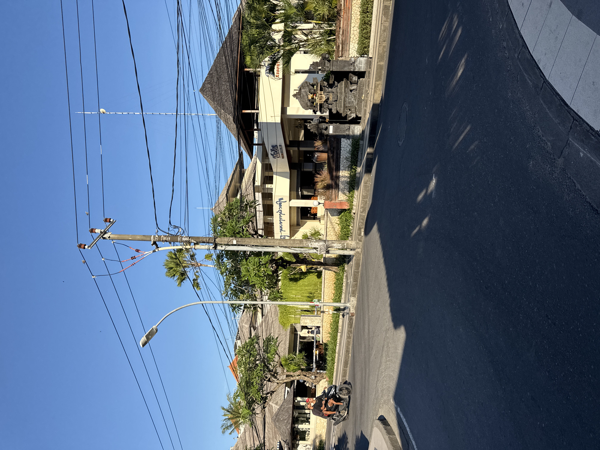 The image shows a street scene in Kuta, Indonesia. A motorcyclist is riding on the right side of the road. A multi-story building with a Balinese-style roofline is on the right side of the street, identified by signage as "Kubu Resort." Across the street are other buildings, also with traditional rooflines. An electric pole with numerous wires is on the left, reaching toward the sky. A street lamp is near the electric pole. The sky is clear and blue. Shadows are cast on the road, indicating a sunny day. The road surface is asphalt.