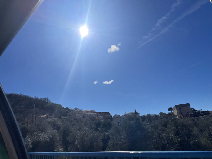 The scene depicts a sunny, clear blue sky over a hilly, vegetated landscape, viewed from a structure resembling a bridge or overpass in Vrijesno, Montenegro. A prominent sun flare is visible in the upper-left quadrant of the sky, which also features two small, wispy white clouds and faint contrails. A blue metal railing occupies the lower portion of the frame, with a dark, curved structural element partially obscuring the far left. The hillsides are densely covered with dark green vegetation, consistent with olive groves. Multiple structures are visible on the slopes: several uncompleted buildings are situated on the left hillside, a cluster of light-colored residential buildings with red-tiled roofs is located in the center-right, and a large, dark, square building, possibly under construction or renovation, stands prominently on the far right hill, flanked by construction vehicles and a solitary tree. No people or active interactions are depicted.