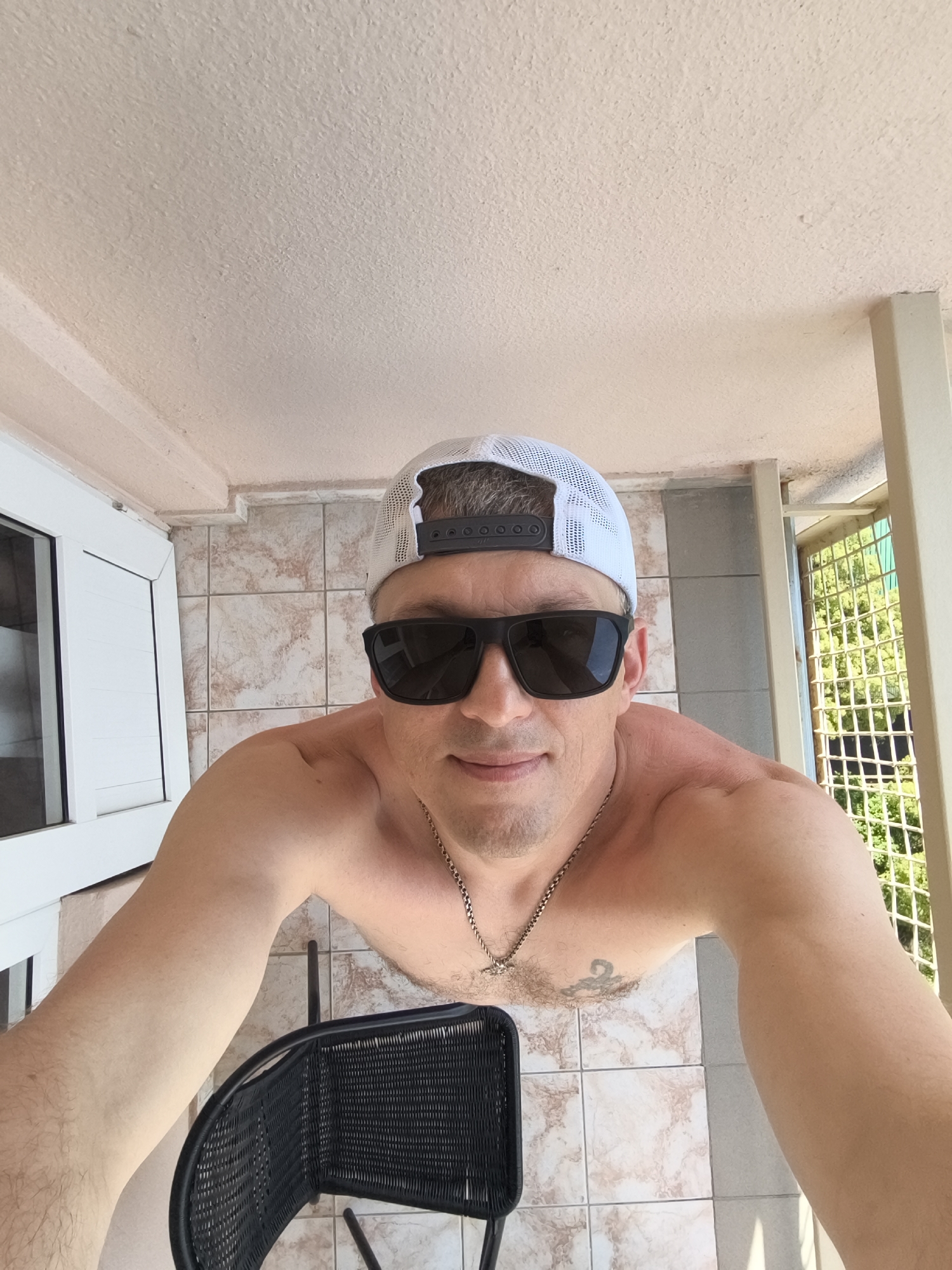 A man wearing a white mesh baseball cap and black sunglasses is captured in a selfie. He is shirtless and appears to be on a balcony or patio, indicated by the tiled floor and a white railing or wall. Behind him, a white window frame is visible on the left, and a metal grate with green foliage beyond it is on the right. A black wicker chair is partially visible in the foreground. The man has a tattoo on his chest and is wearing a silver chain necklace. The lighting suggests it is daytime, likely outdoors. The image does not contain any information to place the scene in Sochi, Russia.