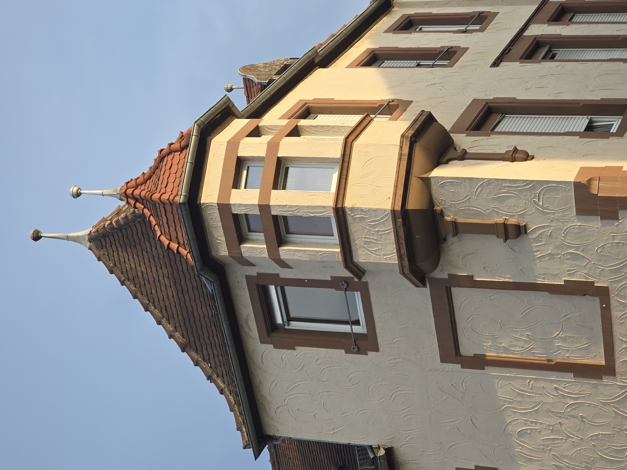 The image shows the corner of a building in Worms, Germany. The building has a textured cream-colored facade with brown trim around the windows and decorative borders. The roof is made of red tiles and has two finials topped with spherical ornaments. There is a bay window on the corner of the building with multiple panes. The sky above is clear and blue. The lighting suggests it is late afternoon or early morning, with long shadows cast on the building. No people or actions are visible in the image.
