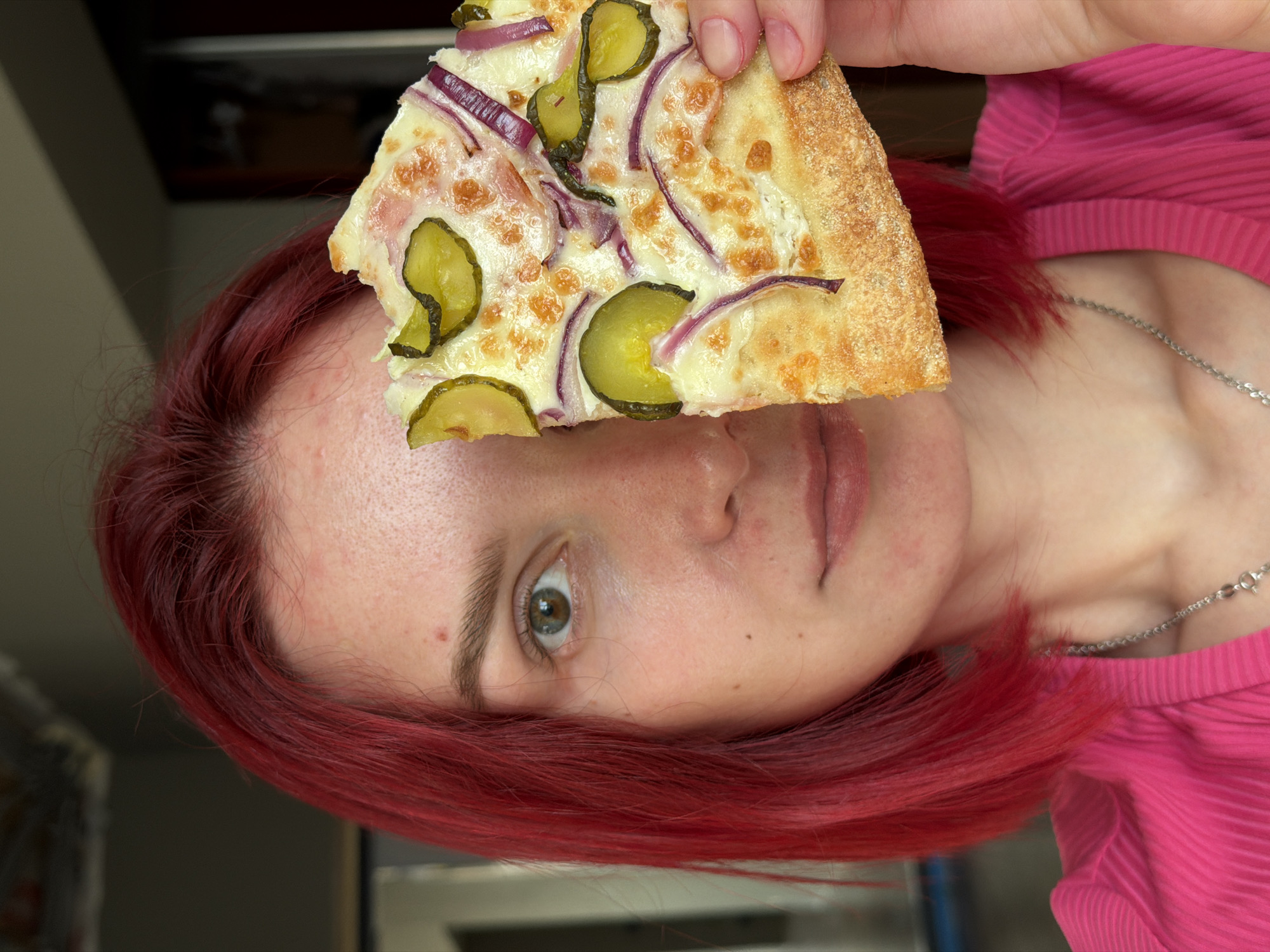 A woman with red hair holds a slice of pizza with toppings including pickles and red onion. The photo is taken indoors. A silver necklace is visible on the woman's neck, and she is wearing a pink shirt. The city and country are not identifiable in the image.