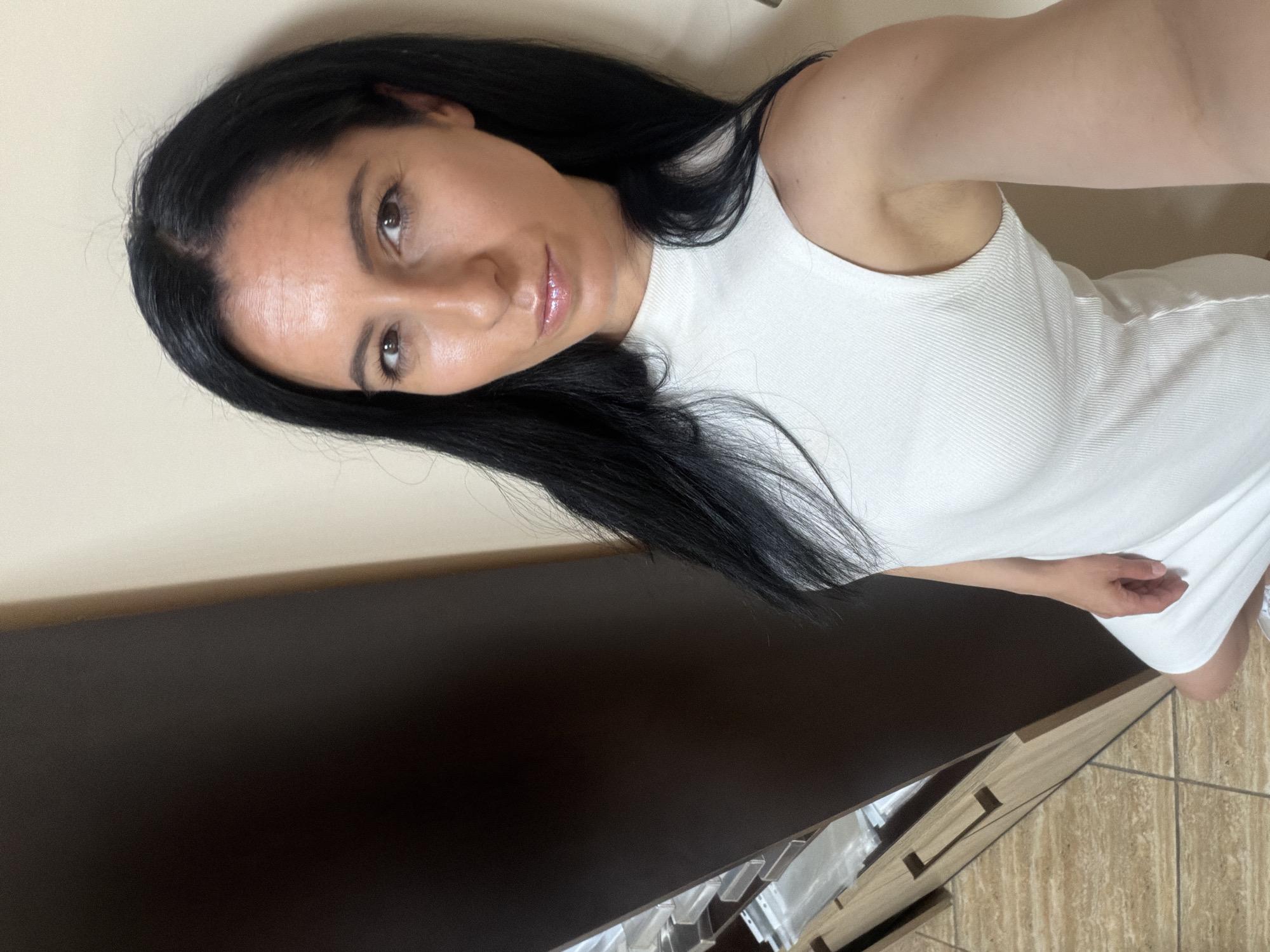 A selfie shows a woman with long, dark, straight hair, dark eyes, and full lips with lip gloss. She is wearing a white, ribbed, high-necked sleeveless top or dress. Her expression is neutral to slightly smiling, and she is looking directly at the camera. Her right arm is visible, extended towards the camera, suggesting she is holding the device.
The setting is indoors, with a plain, light-colored wall forming the upper background. Below this, a dark brown piece of furniture, possibly a dresser or cabinet, spans across the lower part of the image. This furniture has a lighter, possibly wooden, base with visible drawer handles or decorative cut-outs. The floor, visible in the bottom right, is tiled with light brown or beige tiles in a diagonal pattern. The overall lighting is soft, likely from an indoor source, and the general ambiance appears calm. No visible text is present in the image. The image is oriented vertically, but the woman is positioned horizontally, suggesting it was taken and then rotated.