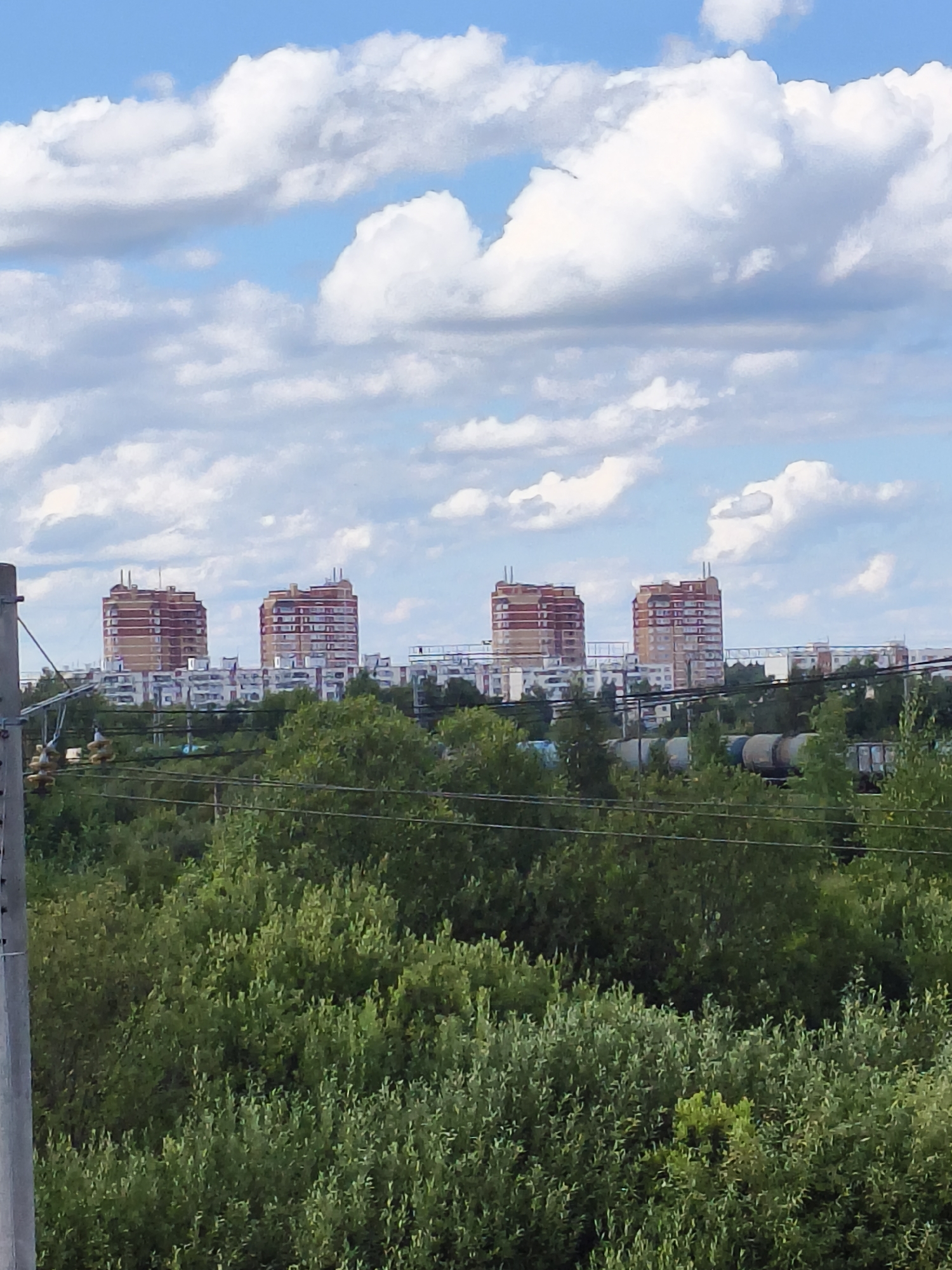 The image captures a view of apartment buildings against a sky filled with cumulus clouds. In the foreground, a dense canopy of green trees dominates the scene, with power lines and a utility pole visible on the left. Behind the trees, several multi-story buildings are arrayed. Four particularly distinct, tall, cylindrical-shaped apartment buildings with reddish-brown facades and horizontal banding stand out in the middle ground. These buildings are spaced apart, and their tops are flat. In front of these taller buildings, a line of lower, more conventional apartment buildings with white and grey facades stretches across the landscape. Further in the background, a train with several carriages is partially visible behind the trees and lower buildings, suggesting the presence of a railway line. The overall environment appears to be a suburban or semi-urban area. The specific location of Kievsky, Russia is not discernible from the visual information alone, as the image lacks any identifying landmarks, signs, or architectural styles that are uniquely indicative of that specific locality.