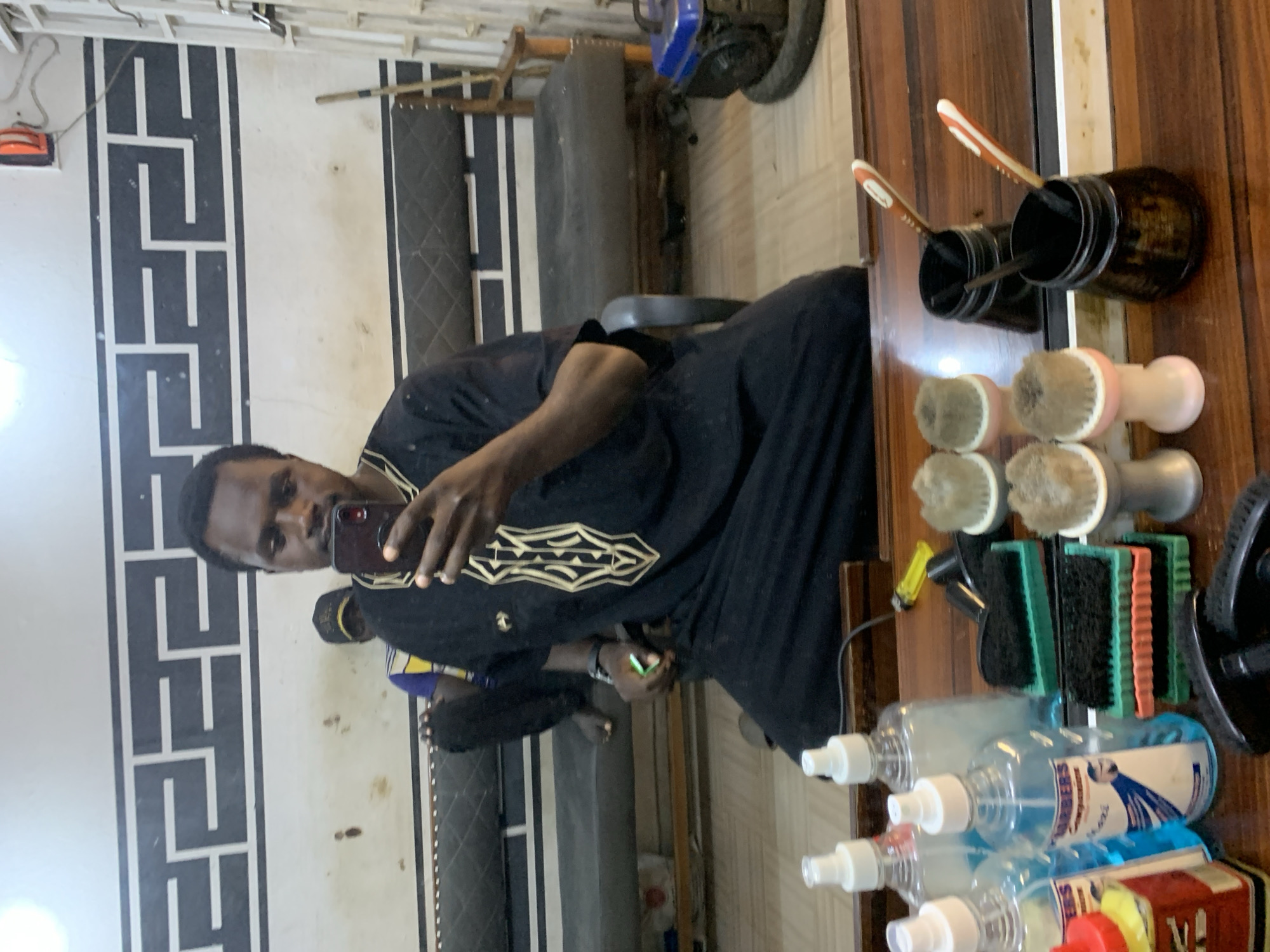 The image depicts a man taking a selfie in a barbershop, likely located in Jos, Nigeria. The man is wearing a traditional black garment with gold embroidery. He is holding a smartphone and appears to be in the process of taking a picture. In the background, another person is seated on a bench. The barbershop features a patterned wallpaper, a sofa, and a mirror reflecting the scene. The foreground of the image shows a wooden counter with various grooming tools, including shaving brushes, combs, and bottles of hair products. A yellow lighter is also visible on the counter. The overall impression is that of a casual setting within a business establishment.