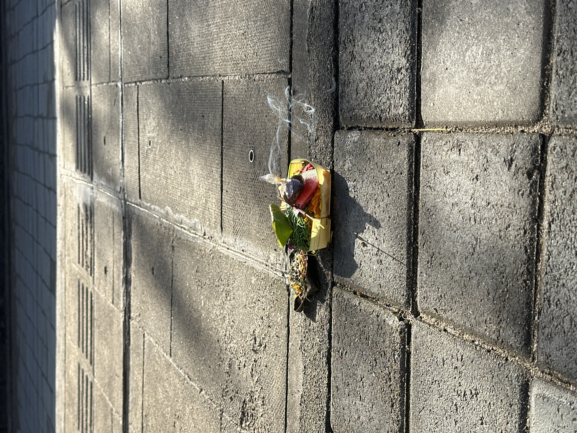 The image shows a Balinese Canang Sari offering placed on a gray brick wall. The offering is in a small woven basket and contains flowers, leaves, rice, fruit, and a smoldering incense stick, producing smoke. The location is in North Kuta, Indonesia.