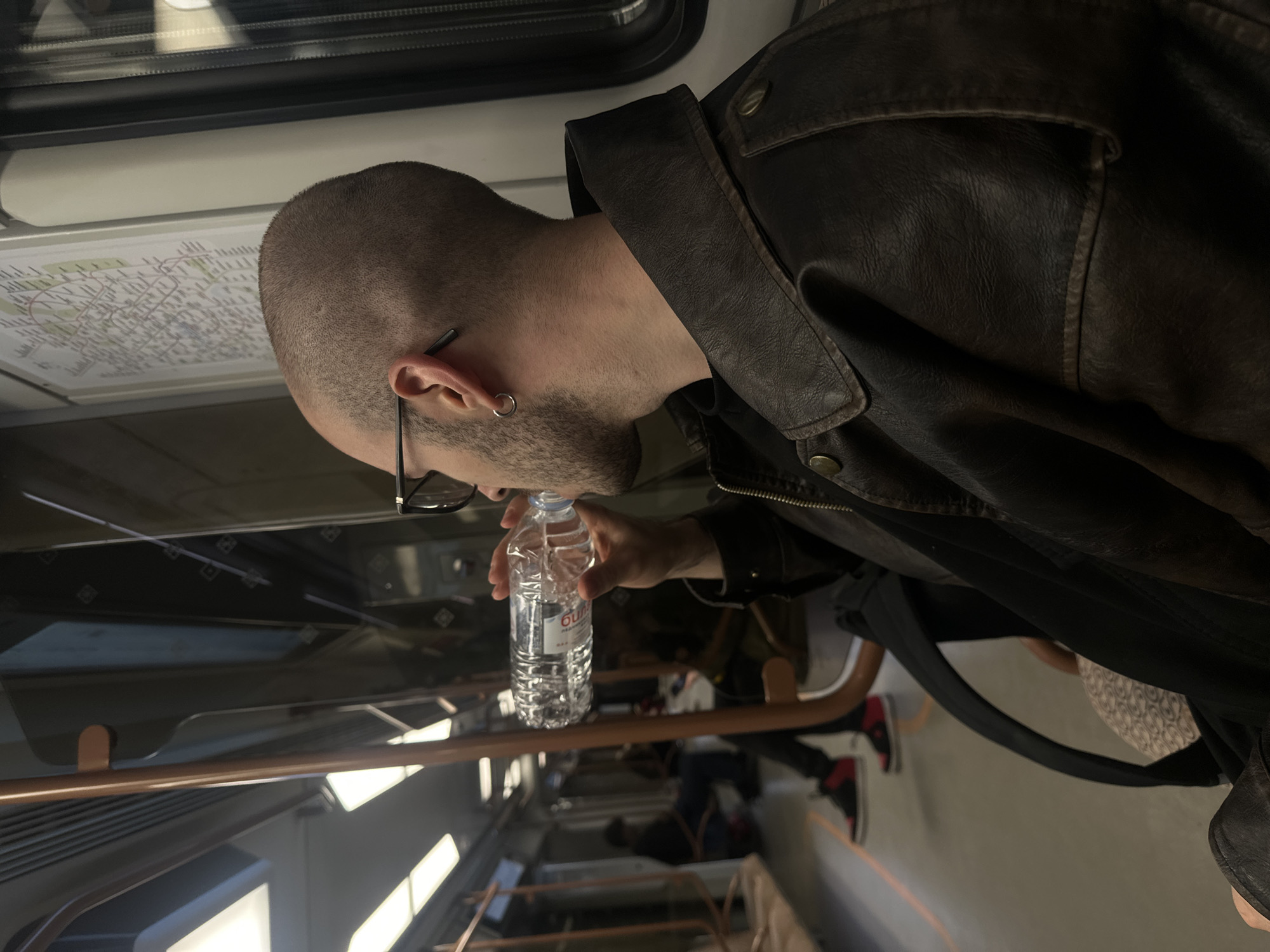 Inside a Moscow, Russia Metro train, a man with a shaved head, wearing glasses and a leather jacket, is drinking from a plastic water bottle. A train route map is visible above him. Seated passengers and handrails are also visible.