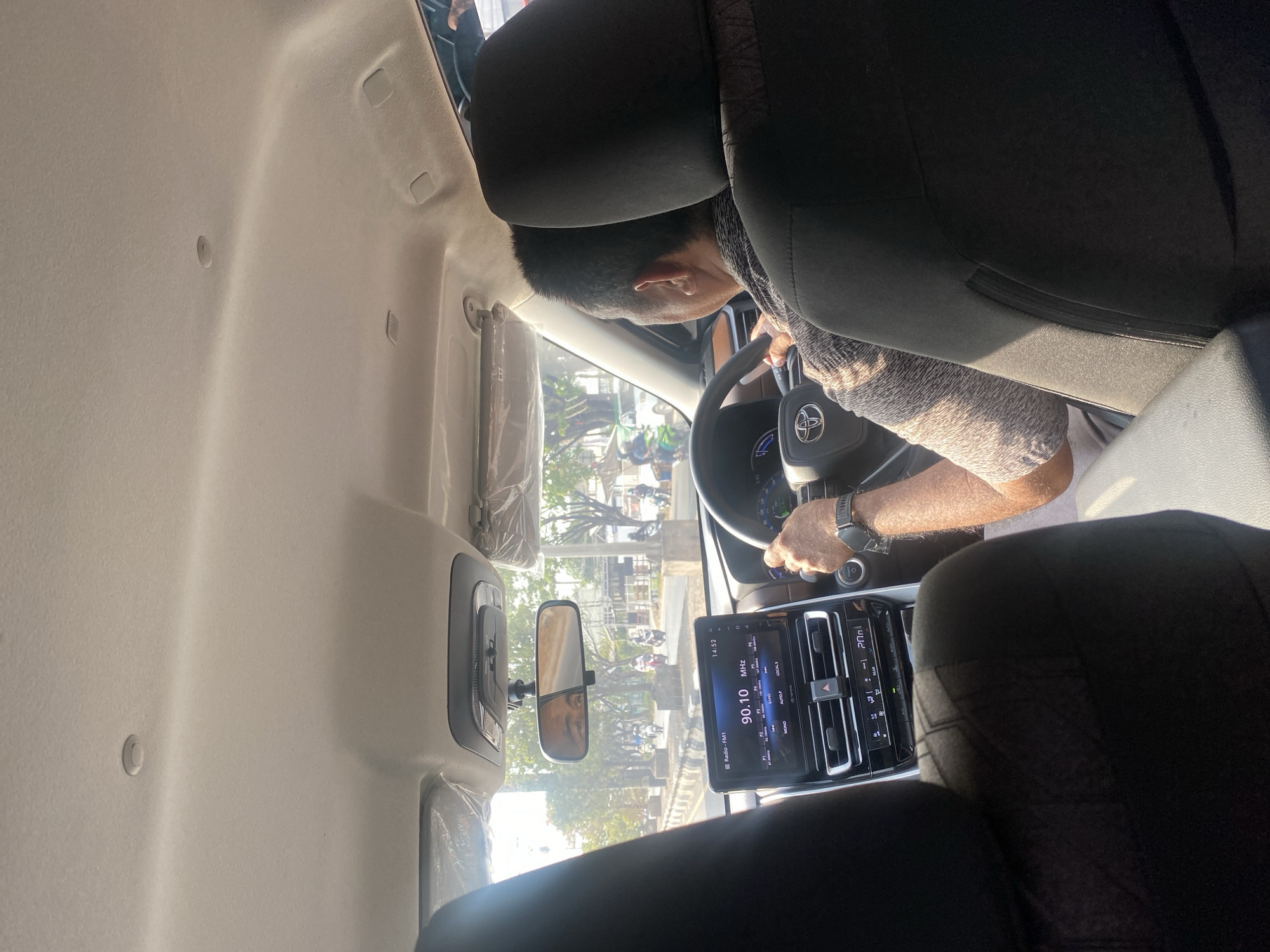The image shows the interior of a car looking forward from the back seat. A man is driving, his hands on the steering wheel. The car's dashboard is visible, featuring a touchscreen infotainment system displaying the radio station "Radio - FM1" and the time "14:52." Sunlight streams in from the windows, illuminating the interior. Outside, a street scene unfolds with trees, other vehicles, and a building. The overall impression is of a daytime drive on a paved road, possibly in a suburban or urban environment. The specific location of Mandar Sari, Indonesia, is not explicitly indicated within the image itself.