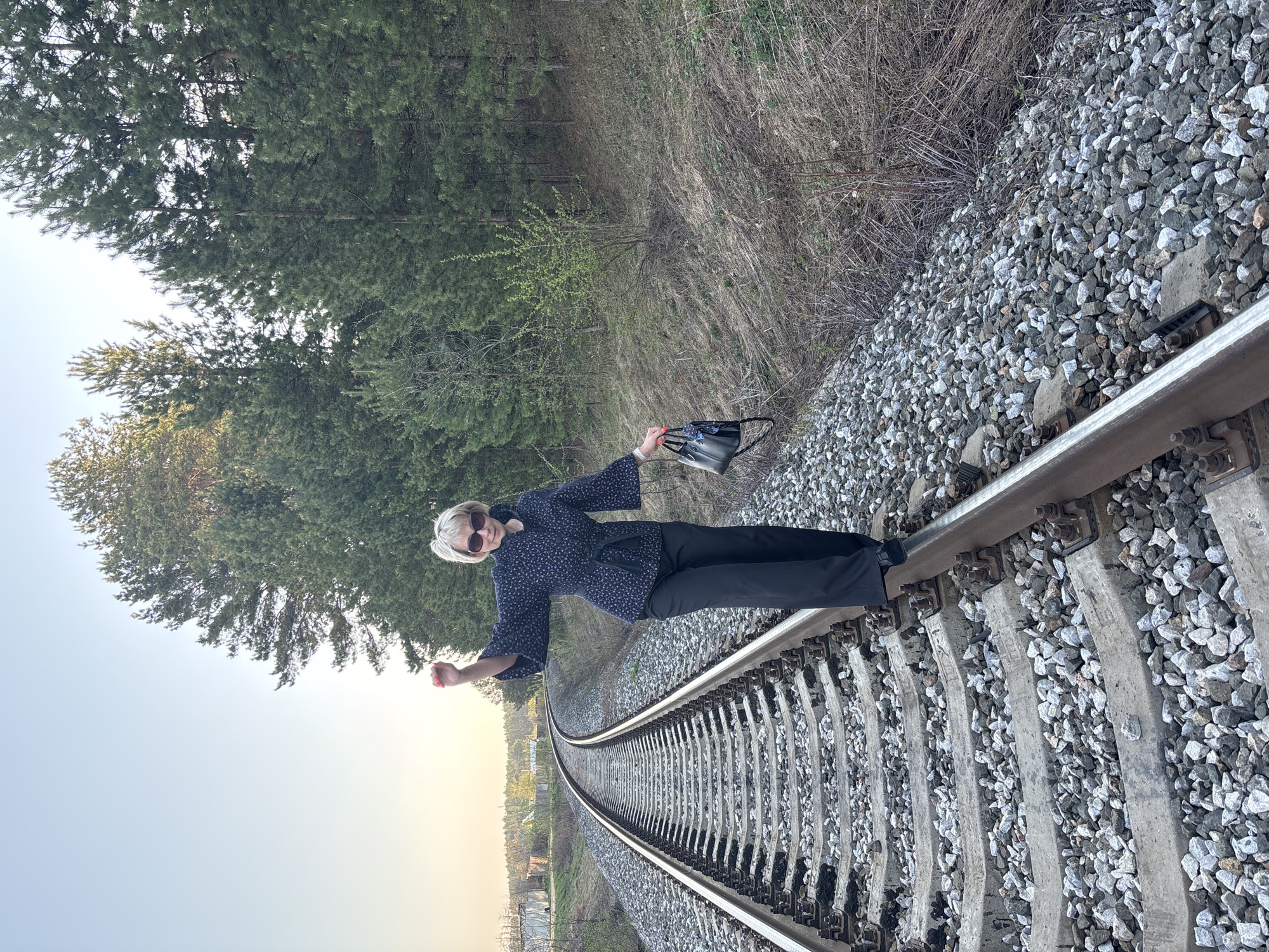A woman stands on a railroad track in Chaikovsky, Russia. She has short blonde hair and wears sunglasses, a dark-colored, patterned blouse, and dark pants. She holds a black purse in one hand and extends the other arm. The railroad track curves into the distance. One side of the track is bordered by trees and brush, while the other shows a more open landscape with a hint of a horizon. Gravel surrounds the tracks.