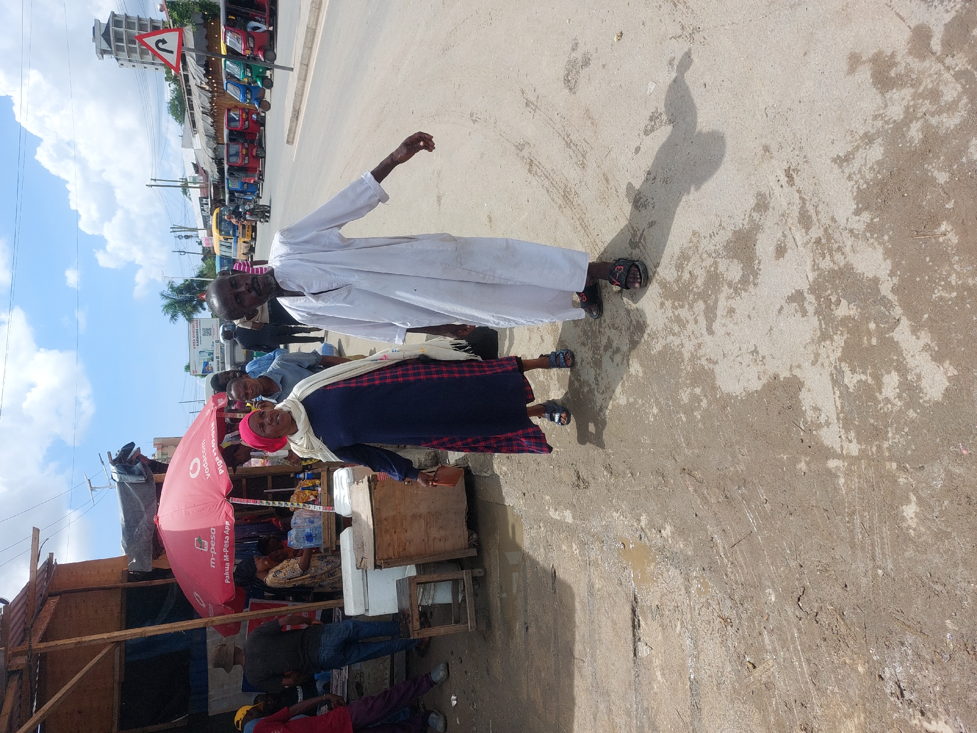 A street scene in Dar es-Salaam, Tanzania, displays a vendor's stall under a Vodafone-branded umbrella, offering various items. An elderly man in a white robe walks beside an elderly woman wearing a blue dress with red plaid and a pink headscarf. They cast long shadows on the ground. In the background, there are several tuk-tuks parked along the roadside, a building with an arrow-shaped road sign, and utility poles with overhead wires. The ground is uneven and possibly muddy.