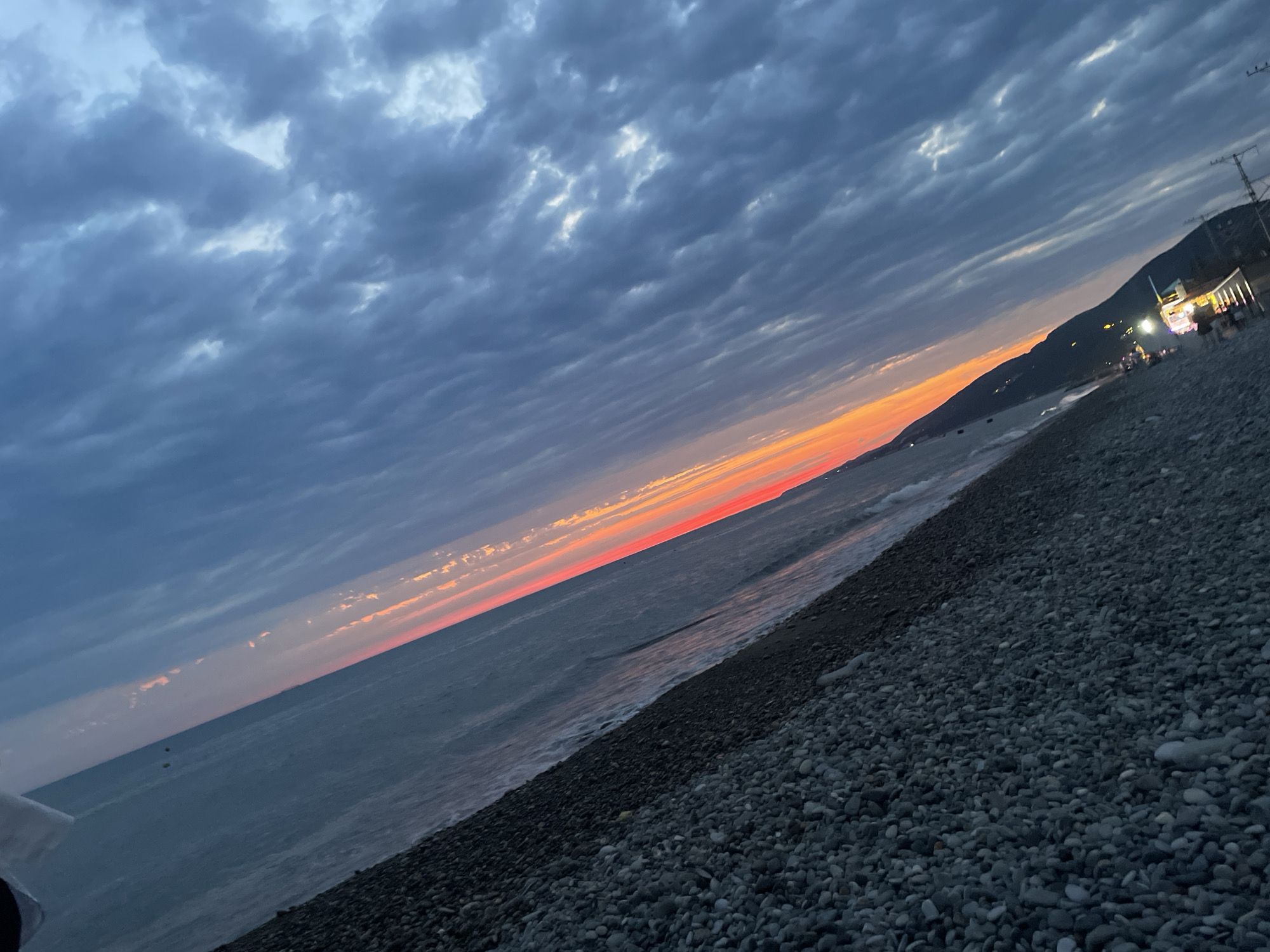 The image depicts a pebble beach at dusk, with the sea on the left and a hillside on the right. The sky is filled with dark, textured clouds, with a vibrant band of orange and red sunset visible near the horizon. A small cluster of lights, possibly indicating buildings or stalls, are illuminated on the hillside. Waves gently lap at the shore. The overall scene is a serene, natural landscape with a touch of human presence.