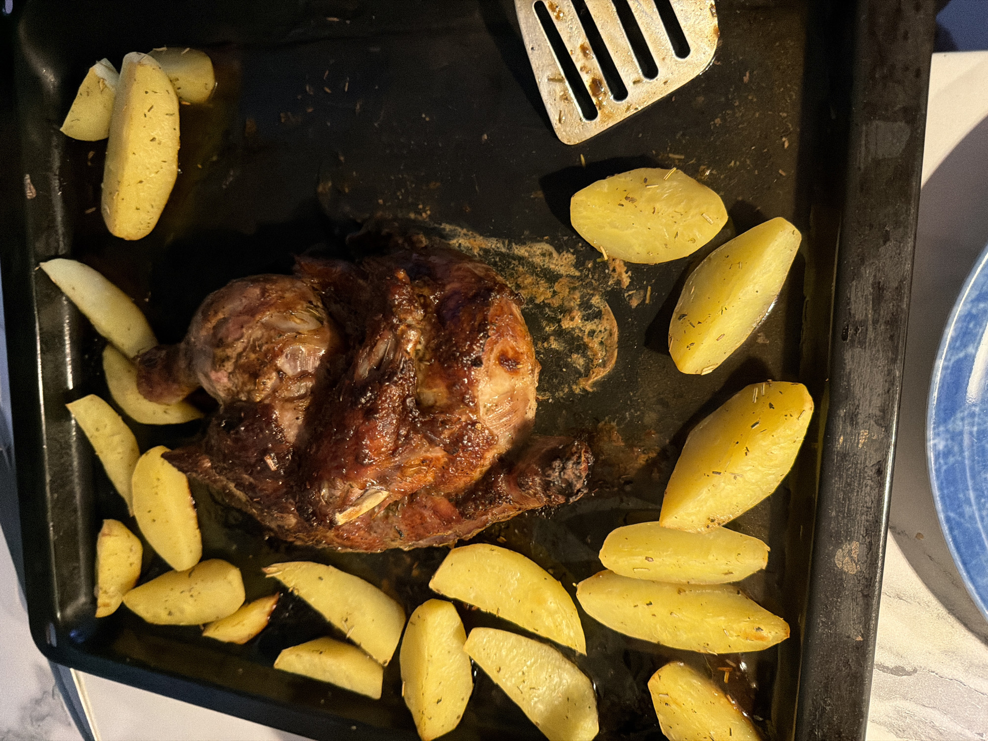 In Novoalexandrovka, Russia, a roasted chicken and potato dish is presented on a dark baking sheet. The chicken, browned and glistening with juices, is positioned centrally. Scattered around the chicken are numerous golden-brown potato wedges, seasoned with herbs. A silver spatula rests on the right side of the baking sheet, near a cluster of potatoes. The baking sheet itself appears used, with some food residue evident. The overall presentation suggests a home-cooked meal.