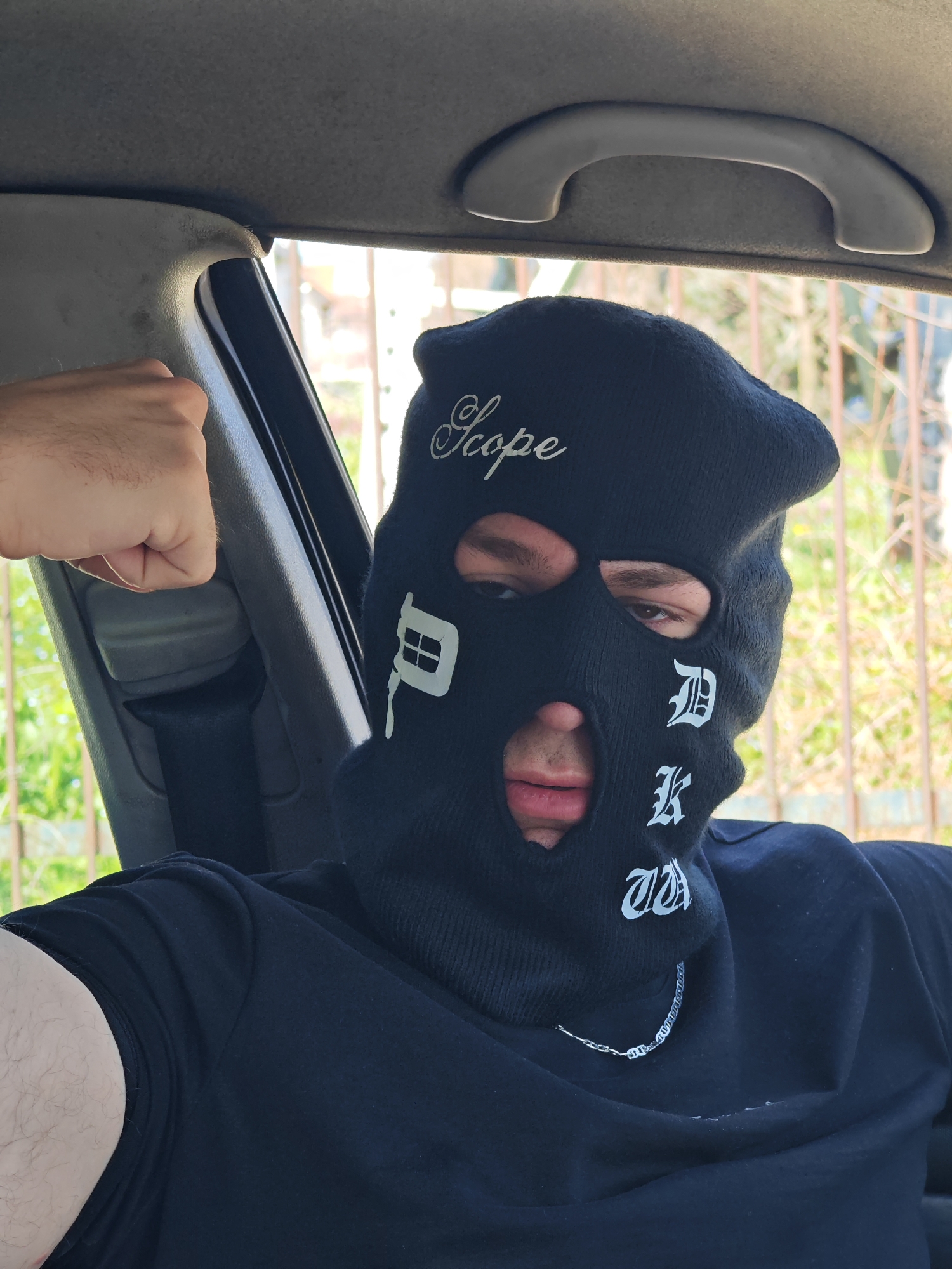 The image presents a single individual seated within the interior of a vehicle, purportedly located in Shumen, Bulgaria. The person is wearing a black balaclava, which covers the entire head and face, revealing only their eyes, eyebrows, and mouth. The balaclava features white stylized lettering: "Scope" written in a script font across the forehead, and a vertical sequence of symbols or letters resembling a building icon or 'P', followed by 'D', 'K', and 'TA' on the wearer's right side of the face. The individual has brown eyes and dark eyebrows, and their lips are visible. They are wearing a black short-sleeved t-shirt, with a silver-colored link chain visible around their neck. The person's left arm (viewer's right) is raised and flexed, with the hand forming a closed fist. The vehicle's interior includes a light grey headliner, an overhead grab handle, and a grey A-pillar trim panel. A dark-colored seatbelt is also visible. Through a window in the background, blurred green foliage and a light-colored, vertically-barred fence or gate are discernible, indicating an outdoor, vegetated environment during daylight hours. The individual is looking directly at the camera.