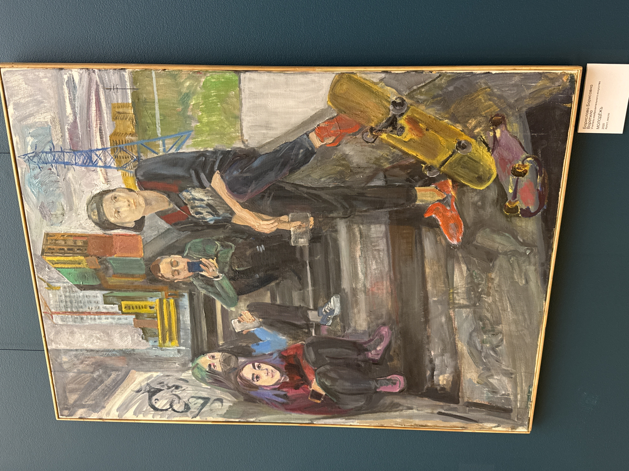 An oil painting depicts a group of young people in a public space. One person is skateboarding, wearing red shoes. Others are sitting or standing, some holding phones, possibly taking photos or videos. The background features buildings and a crane, suggesting an urban construction site. The painting is on display, framed, and hanging on a dark blue wall. The location, based on the label (written in Russian), is Moscow, Russia. The painting style is expressive and somewhat impressionistic, with visible brushstrokes and a focus on capturing the atmosphere of a modern urban scene.