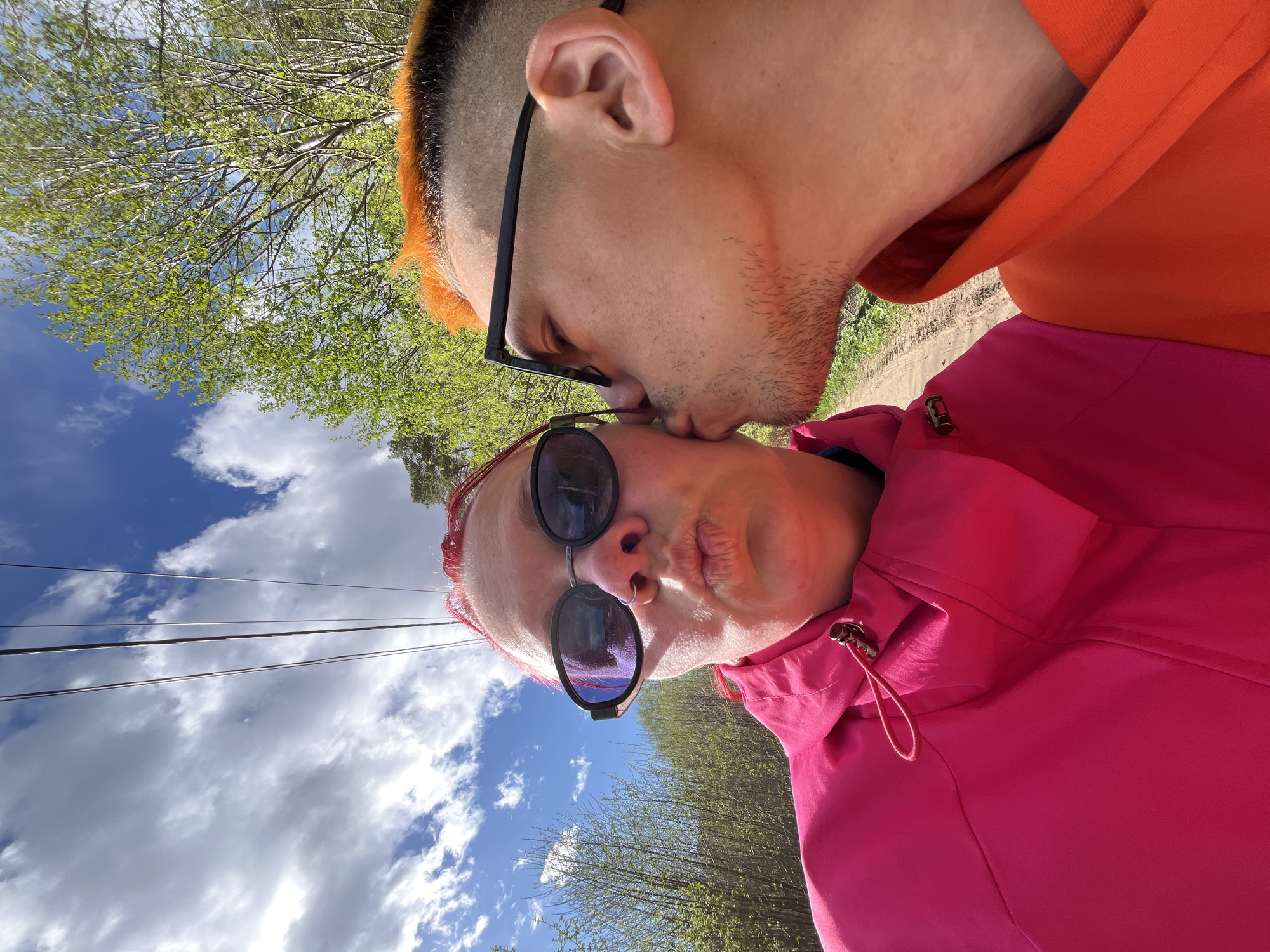 The image shows two people in an outdoor setting. One person with orange hair and black-framed glasses is kissing the cheek of another person wearing sunglasses and a pink jacket. The background features trees, a blue sky with clouds, and power lines. It is not possible to determine the location in the city of Chaikovsky, Russia.
