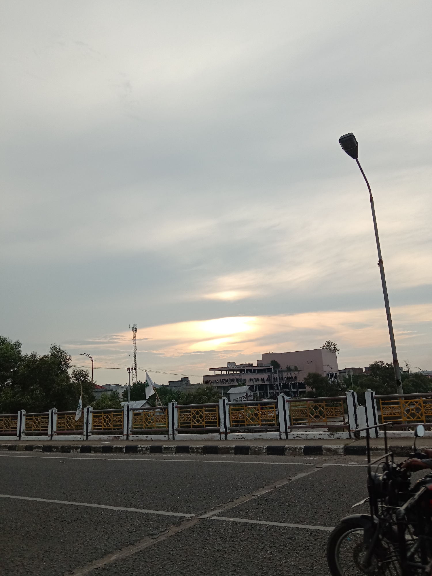 The image shows a street scene at sunset in Lhokseumawe, Indonesia. The viewpoint is from a bridge with a yellow and white decorative railing. A two-lane road is in the foreground with a motorcycle partially visible on the right. In the background, there are buildings, trees, a communication tower, and flags. The sky is cloudy with the sun appearing through a gap. A tall street light stands to the right of the frame.