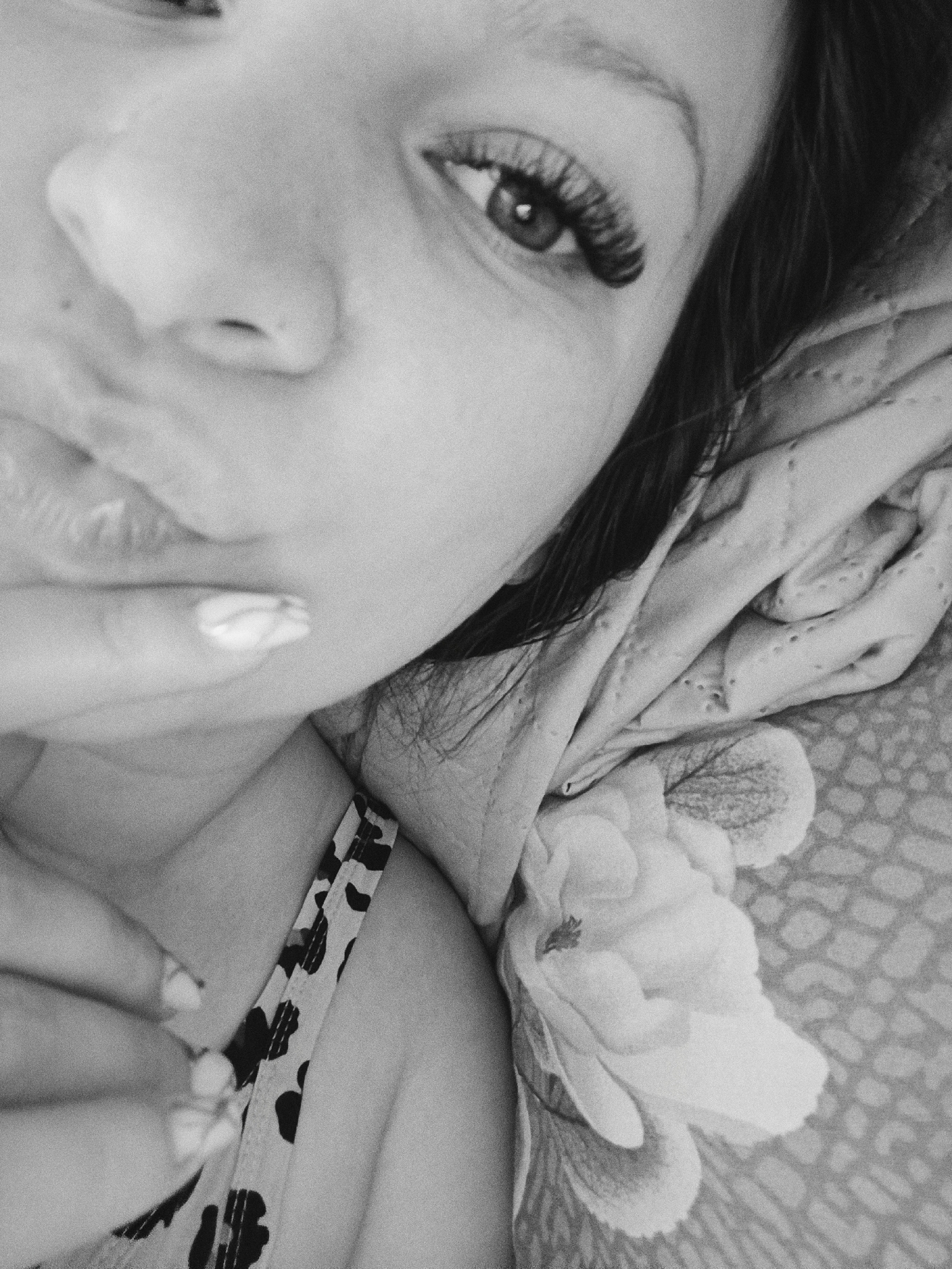 A close-up, grayscale image shows a person with their hand near their mouth. The individual has long eyelashes and appears to be lying down, with a floral-patterned fabric or blanket beneath them. Part of their patterned garment is visible. The specific location in Tobolsk, Russia, is not discernible from the image alone.