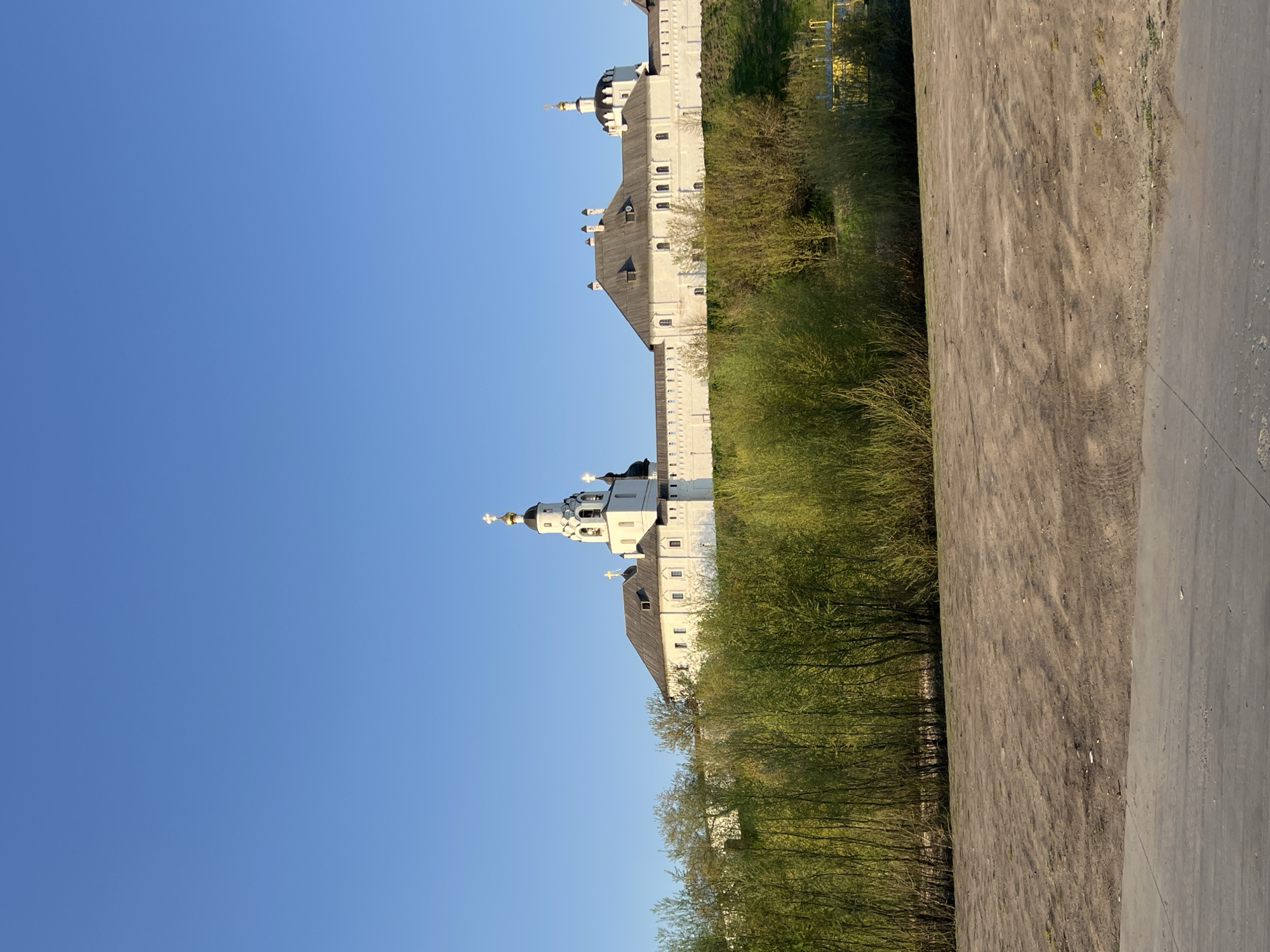 The image features a sideways view of a white walled Orthodox Christian monastery with golden onion domes in Sviyazhsk, Russia. The buildings are partially obscured by trees in the middleground. To the right is a dirt path that leads to a paved road. The sky is clear and blue.