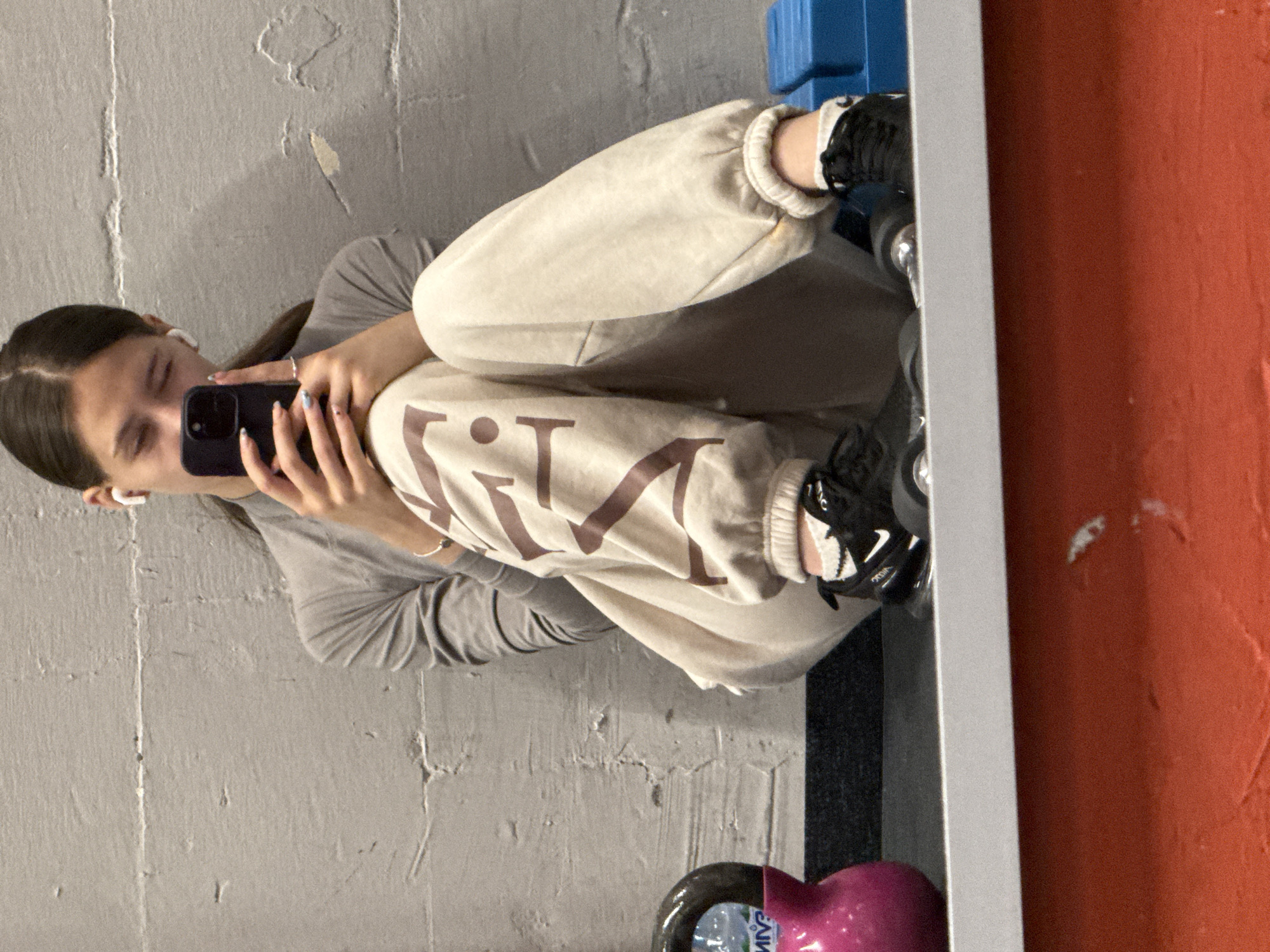The image depicts a young female seated on a dark mat against a textured, light grey concrete wall in a gym setting within Sofia, Bulgaria. She has dark hair styled with a center part and wears white wireless earbuds. Her attire consists of a long-sleeved, light grey mock-neck top and light beige sweatpants featuring large, dark brown vertically printed letters, with "V I N" clearly visible on the left leg. She wears white socks with ruffled tops and black athletic shoes with white brand logos and transparent sole elements. Her fingernails are painted in light pastel colors, and she has a silver ring on her left ring finger and a silver bracelet on her right wrist. The female holds a dark-colored smartphone with a prominent camera module in both hands, positioned in front of her face as if viewing or photographing. Her eyes appear partially closed. To the left, a glossy pink kettlebell with a black handle rests on the mat; its surface reflects partial green and blue text. To the right, stacked blue aerobic steps are visible. The scene is observed through a mirror, with a grey horizontal surface marking the mirror's base. Below this line, a textured red-orange floor is reflected.