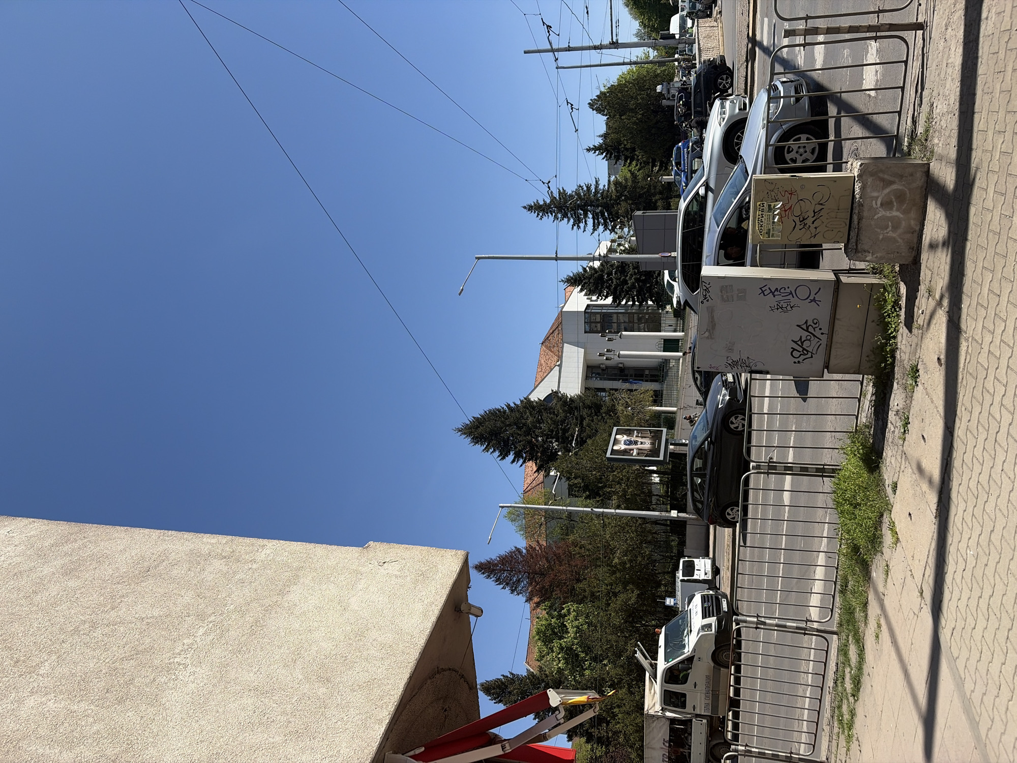 The image captures a sunny, daytime street scene in Sofia, Bulgaria, characterized by urban infrastructure and parked vehicles. The sky is a clear, bright blue, crisscrossed by numerous overhead lines, indicative of a trolleybus or tram system.
On the left, a building with a light-colored, textured facade is partially visible. Further down the street, another building with a reddish-brown roof and white walls can be seen, surrounded by various trees, including tall evergreen conifers and deciduous trees.
The right side of the image features a patterned paved sidewalk, separated from the street by metal barriers. Several utility boxes or electrical cabinets are positioned along the sidewalk, some covered in graffiti. One box has partially legible Cyrillic text, possibly "ОКРИВКА". The street itself is lined with parked cars, including a prominent white utility van with Cyrillic text on its side that reads "ТЕЛКИ СИГНАЛИЗАЦИЯ" (likely "Wires Signaling" or "Cable Signaling"), and a silver SUV.
Bright sunlight casts strong, defined shadows from the barriers and other street furniture onto the sidewalk, suggesting it is mid-day. A tall streetlamp stands prominently, and a billboard-like sign, possibly for watches, is visible in the background behind some trees. The scene depicts a typical, active urban environment with a mix of residential/commercial buildings, green spaces, and transportation infrastructure.