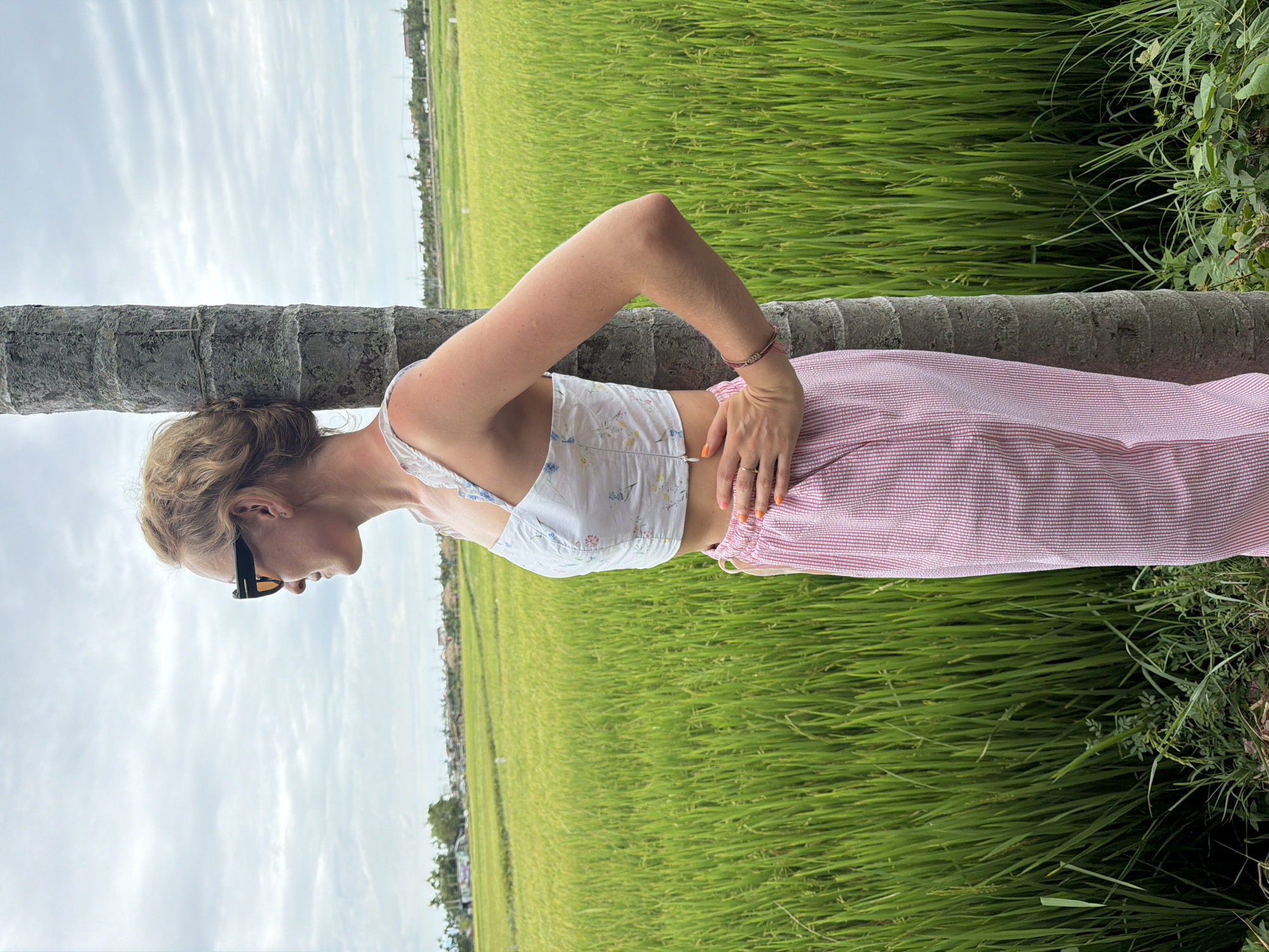 A young woman is pictured leaning against a palm tree in a verdant rice paddy field in Hoi An, Vietnam. She is wearing a white floral print crop top with ruffled straps and loose pink and white striped pants. Her blonde hair is pulled back, and she has on black-framed sunglasses. Her right hand rests on her hip, and her left arm is bent, with her hand near her waist. She is looking off to her left, towards the horizon. The sky is overcast with scattered clouds. The rice field stretches out behind her, a vast expanse of green stalks. In the distance, a few scattered buildings and trees are visible. The overall scene is one of rural tranquility under a cloudy sky.