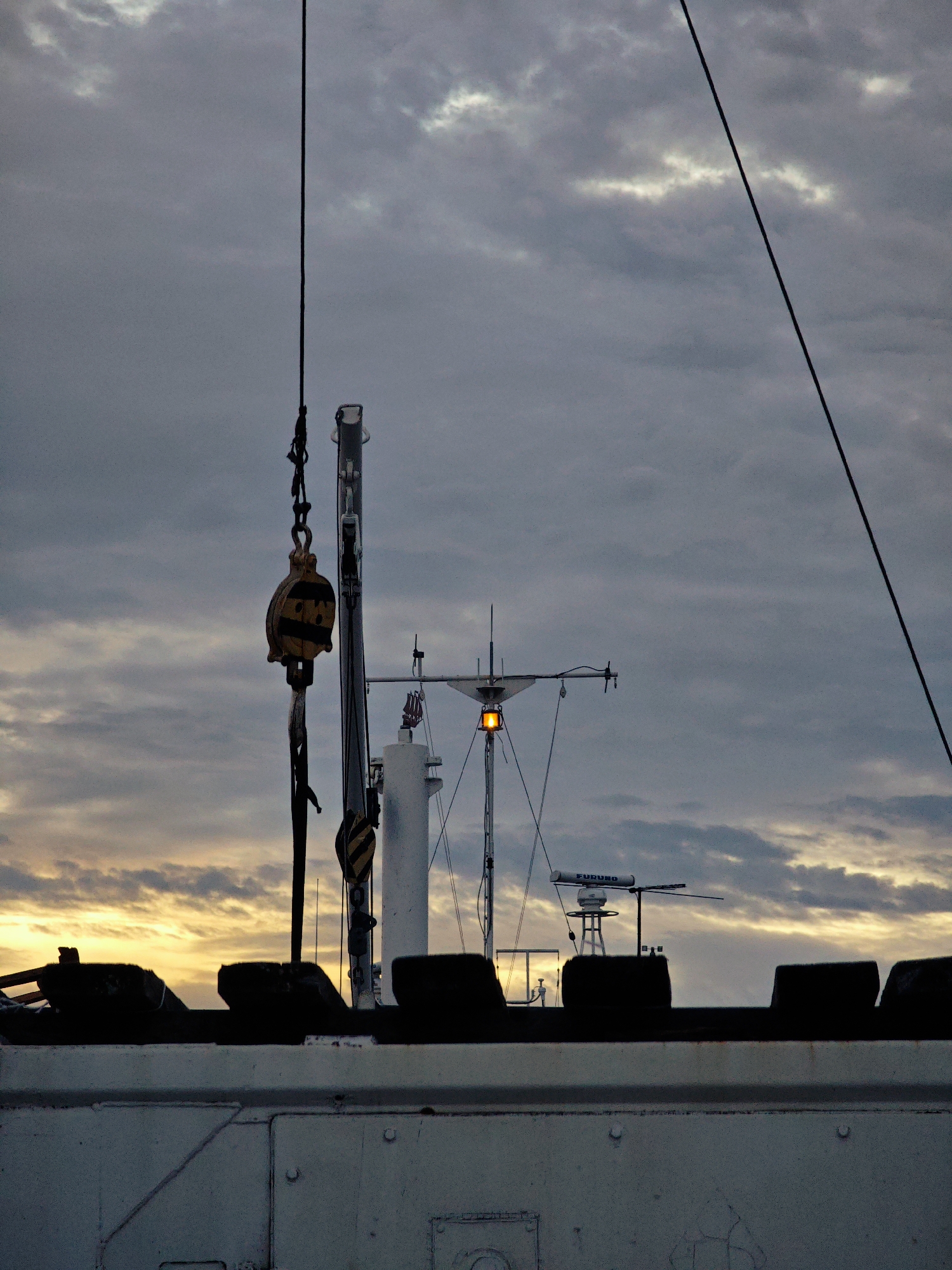 This media file shows the top of a boat with a crane and its pulley system against a cloudy sky at sunset or sunrise. The sky is dramatic with streaks of yellow and orange from the low sun visible through the gray clouds. A tall mast with a bright orange light, possibly a navigation light, is prominent in the center. A radar unit with "FURUNO" written on it is mounted on a shorter mast to the right of the main mast. The boat itself is white and appears to be an older vessel, with visible rivets and some signs of wear on the hull. Dark, silhouetted objects, possibly cargo or equipment, are visible along the railing of the boat. The overall mood is somber and industrial, with a touch of the natural beauty of the sky at twilight. The scene likely takes place at sea or in a port, given the presence of the boat and maritime equipment. No people are visible in the frame.