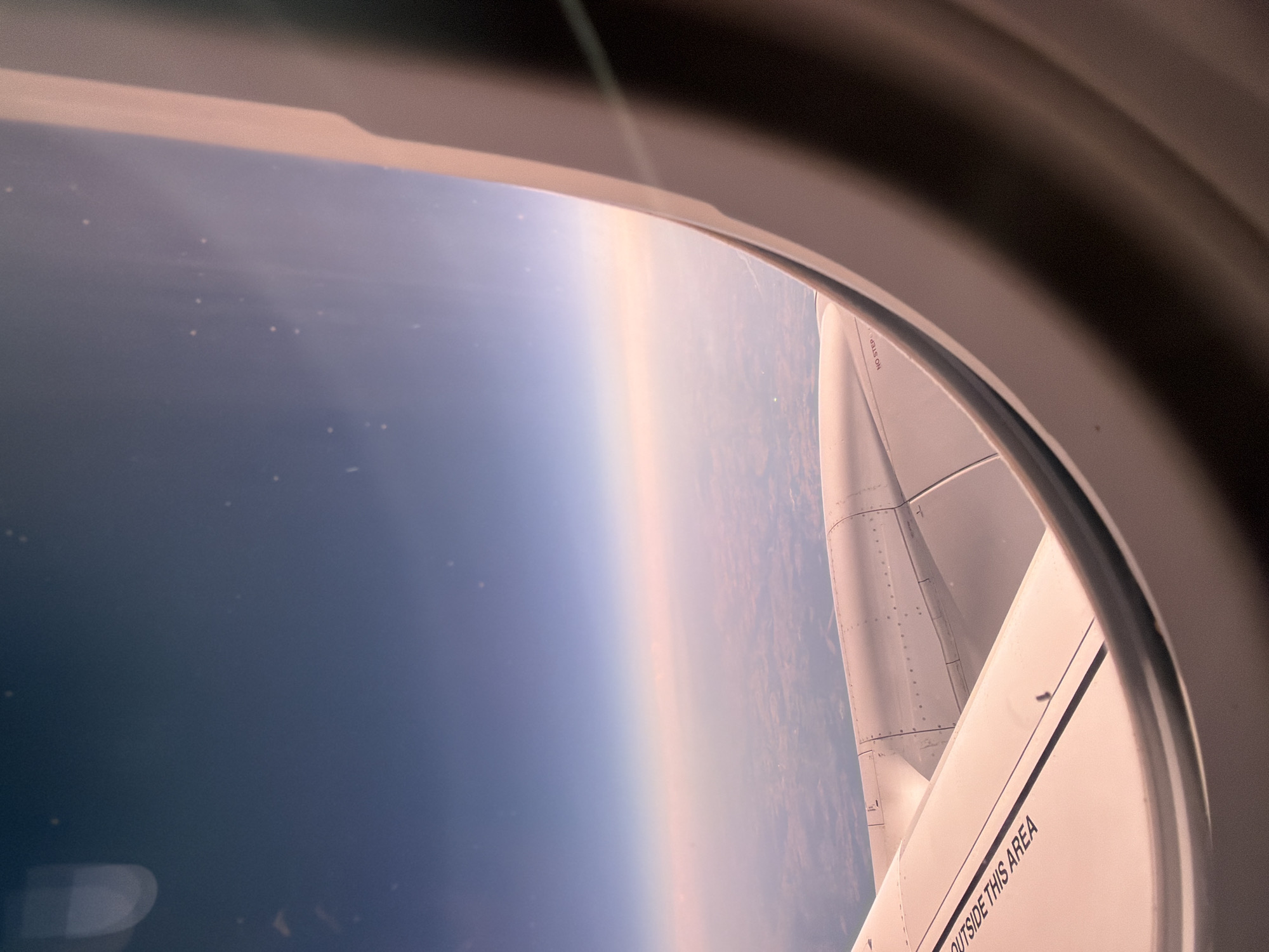 An aerial view from an airplane window shows the wing and engine cowling against a hazy sky. The horizon line is visible, with a gradient of colors from deep blue at the top to pink and orange closer to the ground. The ground below appears to be a patchwork of fields and possibly some structures, suggesting a rural or suburban landscape. The window frame is visible in the foreground, curving around the edge of the image. Text indicating "OUTSIDE THIS AREA" is visible on the wing. The image captures a serene moment during a flight, with the soft lighting indicating either sunrise or sunset. The location in Wolferborn, Germany, is not directly depicted but is the assumed geographic context for this aerial view.