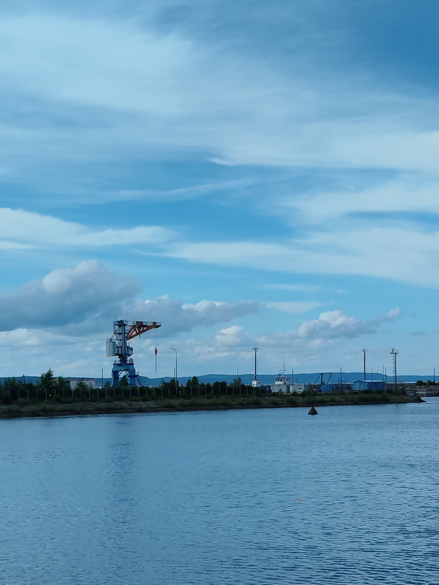 The image depicts a wide, calm body of water in the foreground. In the background, a shoreline is visible, lined with trees and a fence. Several industrial structures are present, including a large blue and orange crane, various buildings, and tall poles with lights. A small boat or structure is situated on the shore near the poles. A red buoy floats in the water. The sky above is a clear blue with scattered white clouds, suggesting it is daytime. The overall scene appears to be an industrial port or waterfront area. There is no visible text in the image.