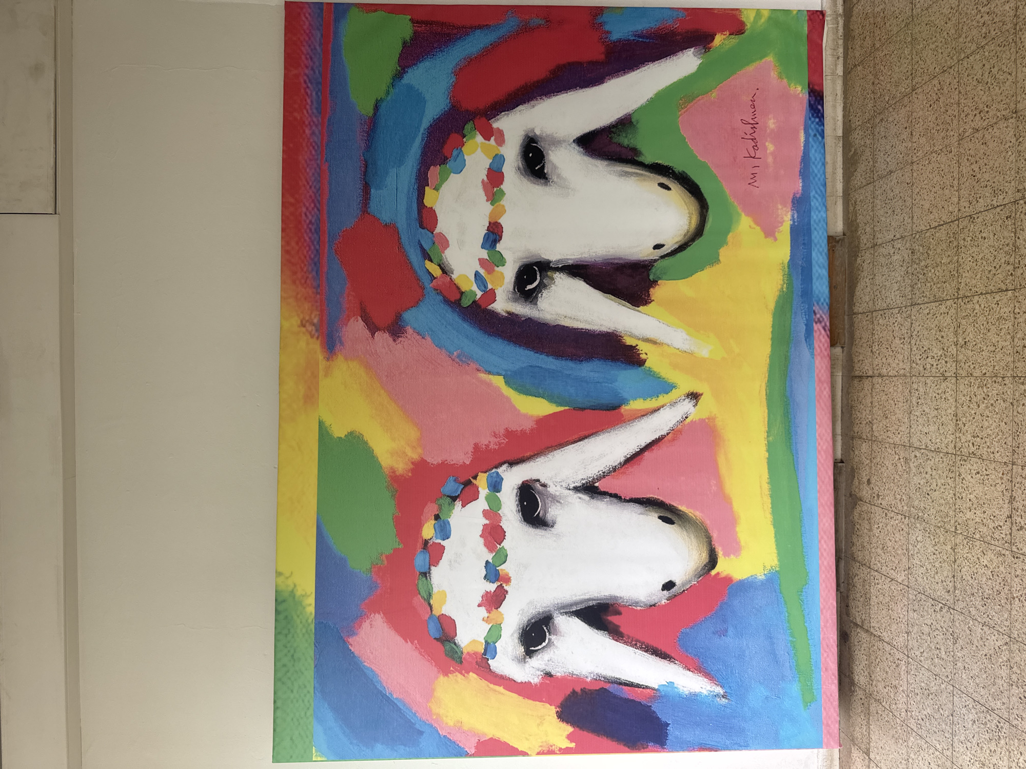 A horizontally-oriented photograph captures an abstract painting hung on a wall, potentially indoors. The artwork features two upside-down white animal heads, possibly donkeys or rabbits, adorned with colorful garlands. The background consists of vibrant brushstrokes of various colors, including blue, red, green, pink, and yellow. A signature is visible in the upper right corner of the painting.
The wall to the left of the painting is painted white, while the wall to the right is covered in beige tiles. It is alleged that this image was taken in the city of Netanya, Israel. However, there is nothing in the image to confirm this statement.