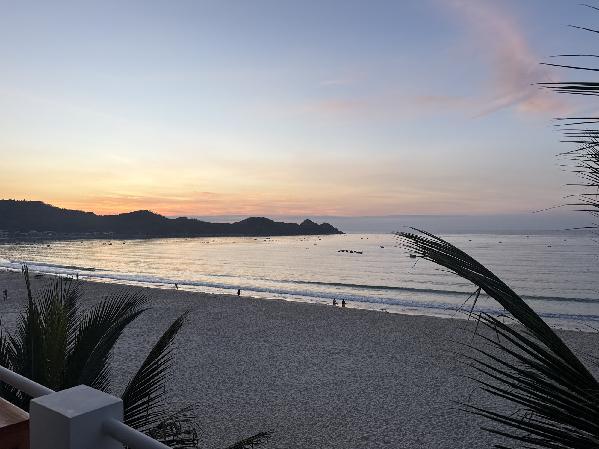 The image shows a serene beach scene at dawn. The sky transitions from a pale blue at the top to warm hues of orange and pink near the horizon, where a silhouetted mountain range is visible. The calm sea reflects the colors of the sky, and gentle waves lap against a sandy beach. A few distant boats dot the water. On the beach, several figures are scattered, some walking along the shore, others near the water's edge. In the foreground, the fronds of a palm tree obscure parts of the view, and a white railing and pillar suggest a balcony or viewing platform. The overall atmosphere is peaceful and tranquil.