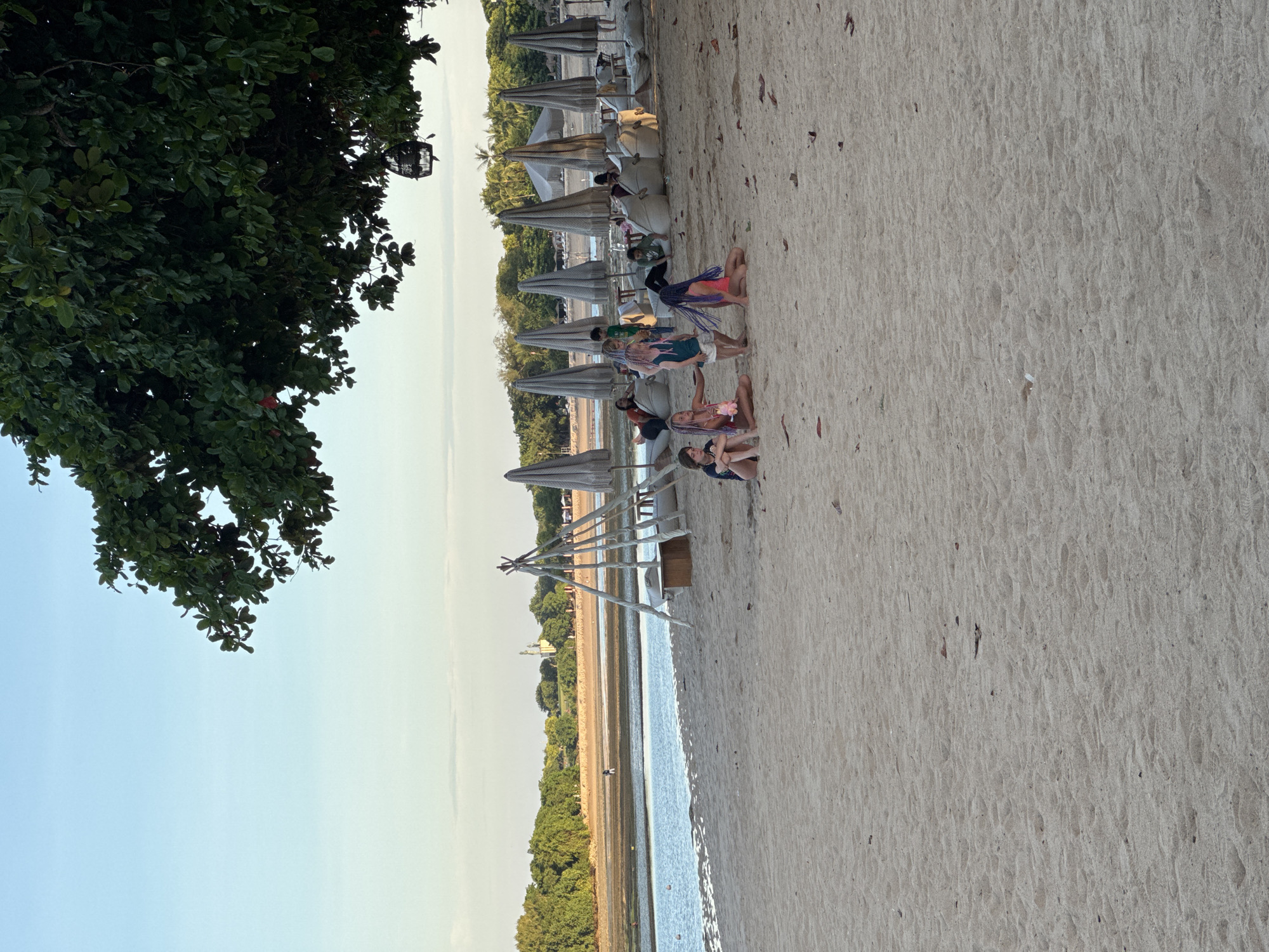 Here's a detailed description of the image:
**Location:** Unknown, Unknown, based on the visual elements, the location is a sandy beach.
**Environment:** The image depicts a beach scene. The sky is clear and bright, indicating daylight. The sand is light-colored and dry, stretching out toward the water. A strip of green vegetation (likely trees and shrubs) runs along the edge of the water, providing a natural boundary.
**Objects:** There are several rows of thatched umbrellas with beach chairs underneath, suggesting a resort or recreational area. A wooden structure with ropes (possibly a lifeguard stand or decorative element) is present.
**People:** A group of people, possibly tourists or vacationers, are sitting and relaxing on the sand near the umbrellas and chairs.
**Actions/Interactions:** The people are engaged in leisurely activities such as sitting, chatting, and enjoying the beach. There's no visible active recreation (e.g., swimming or sports) in the frame.
